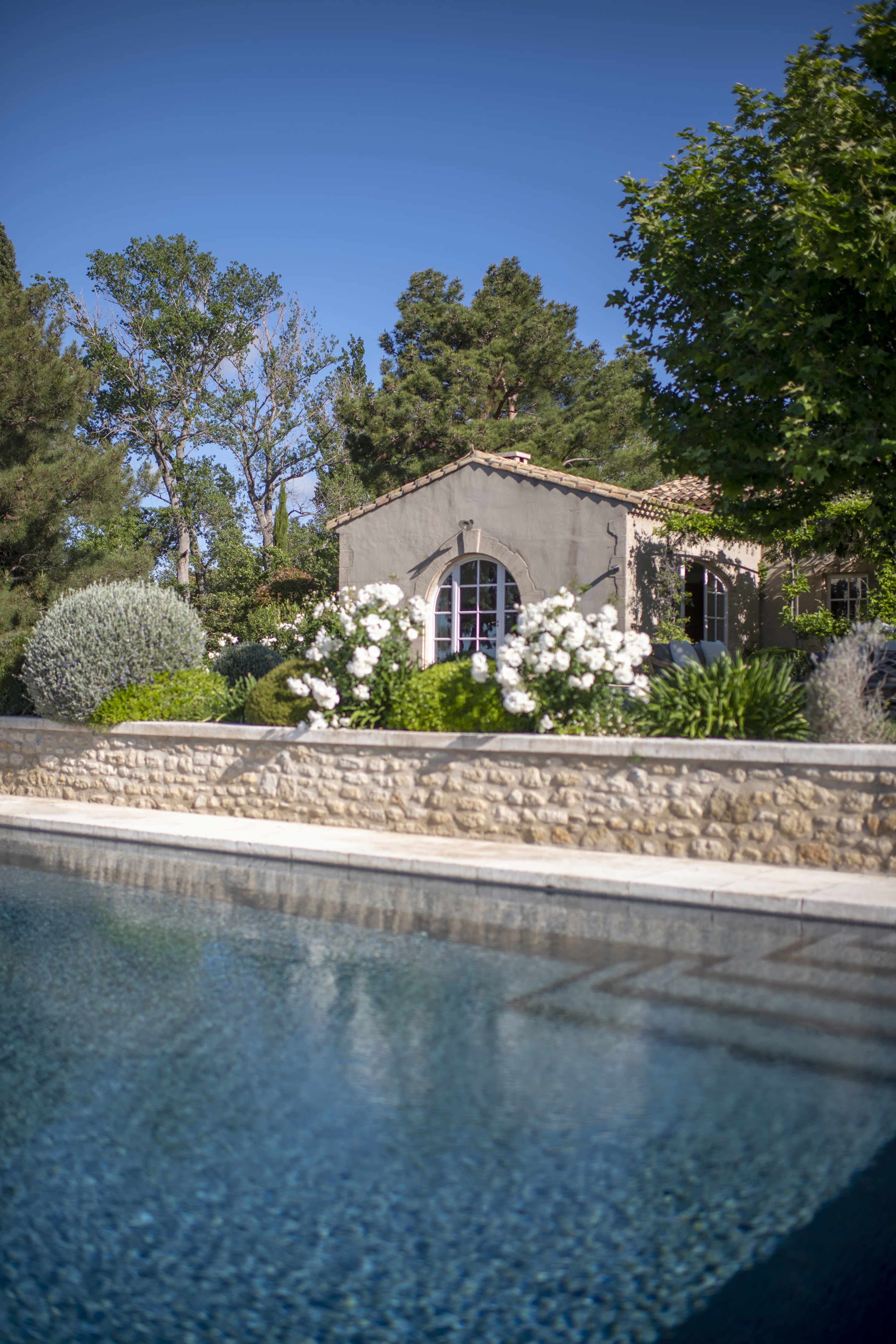 A swimming pool reflects a stone-walled house surrounded by lush greenery and blooming white flowers.