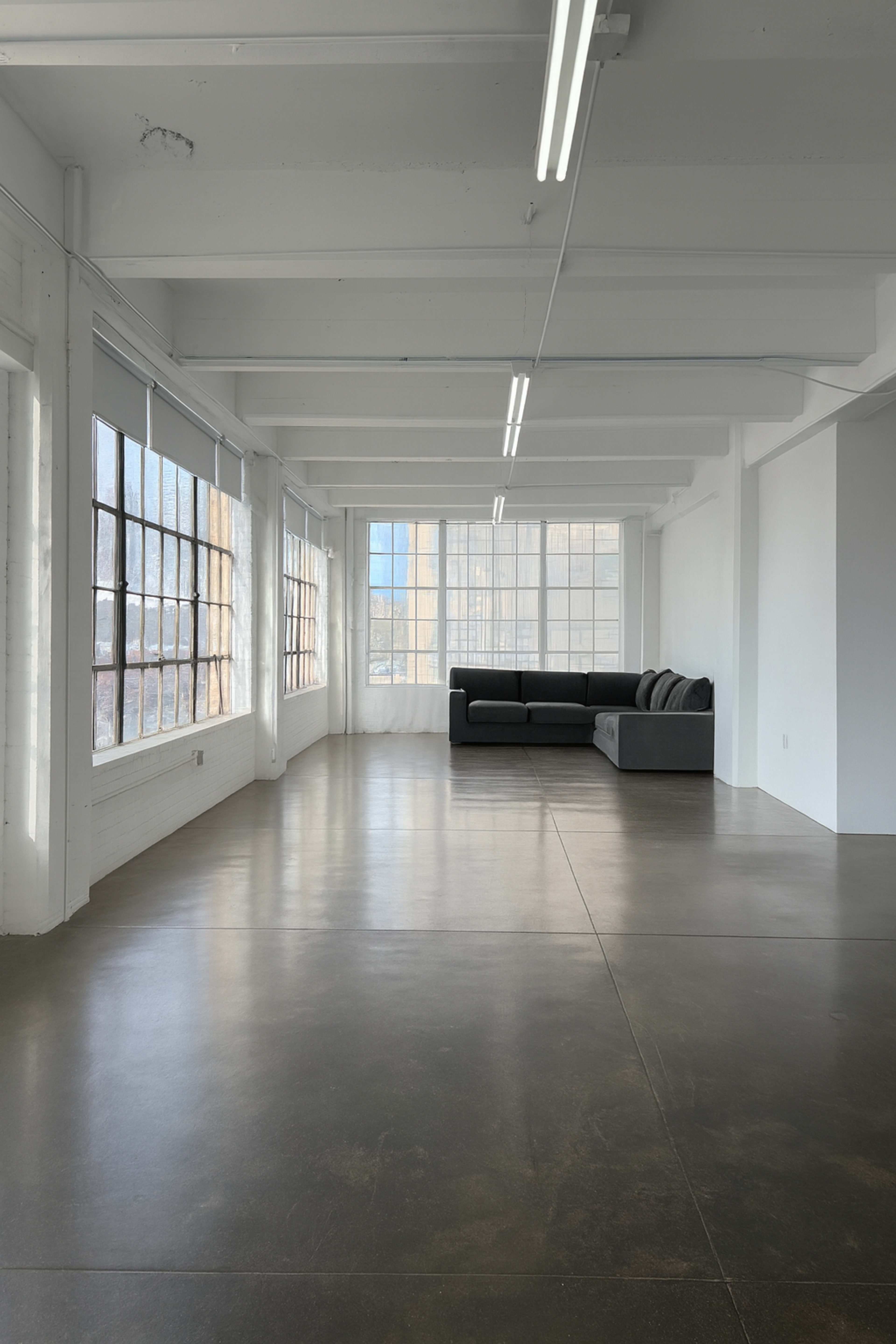 The image shows an empty, well-lit interior space featuring large windows and a gray sectional sofa positioned against one wall.