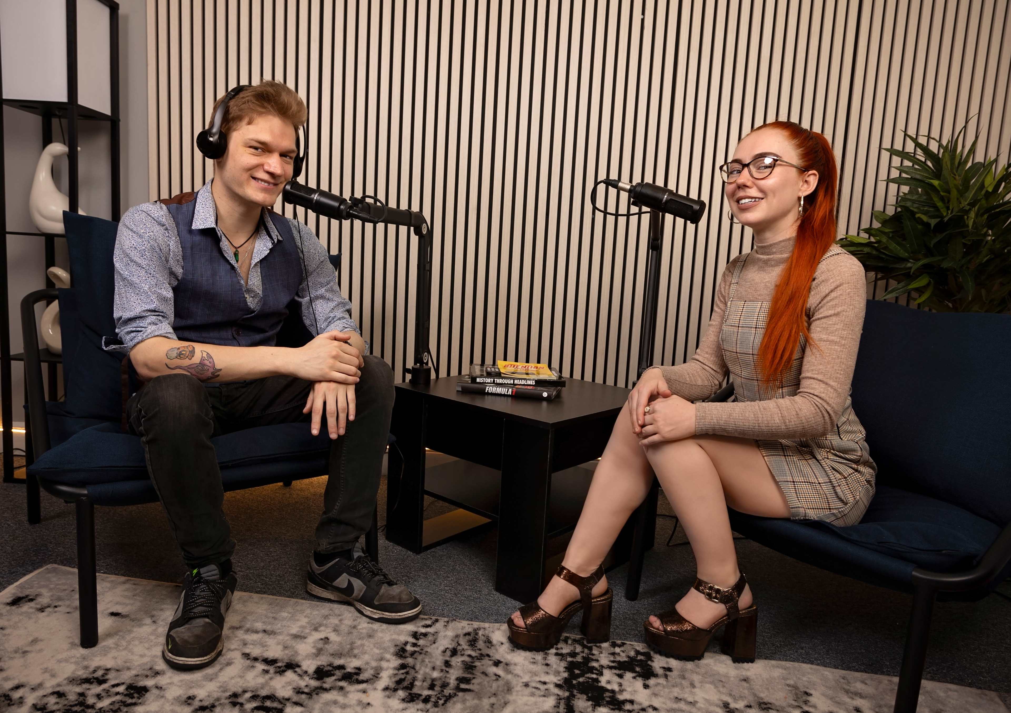 A man and a woman sit in a modern studio with microphones on stands between them, preparing for a podcast recording.