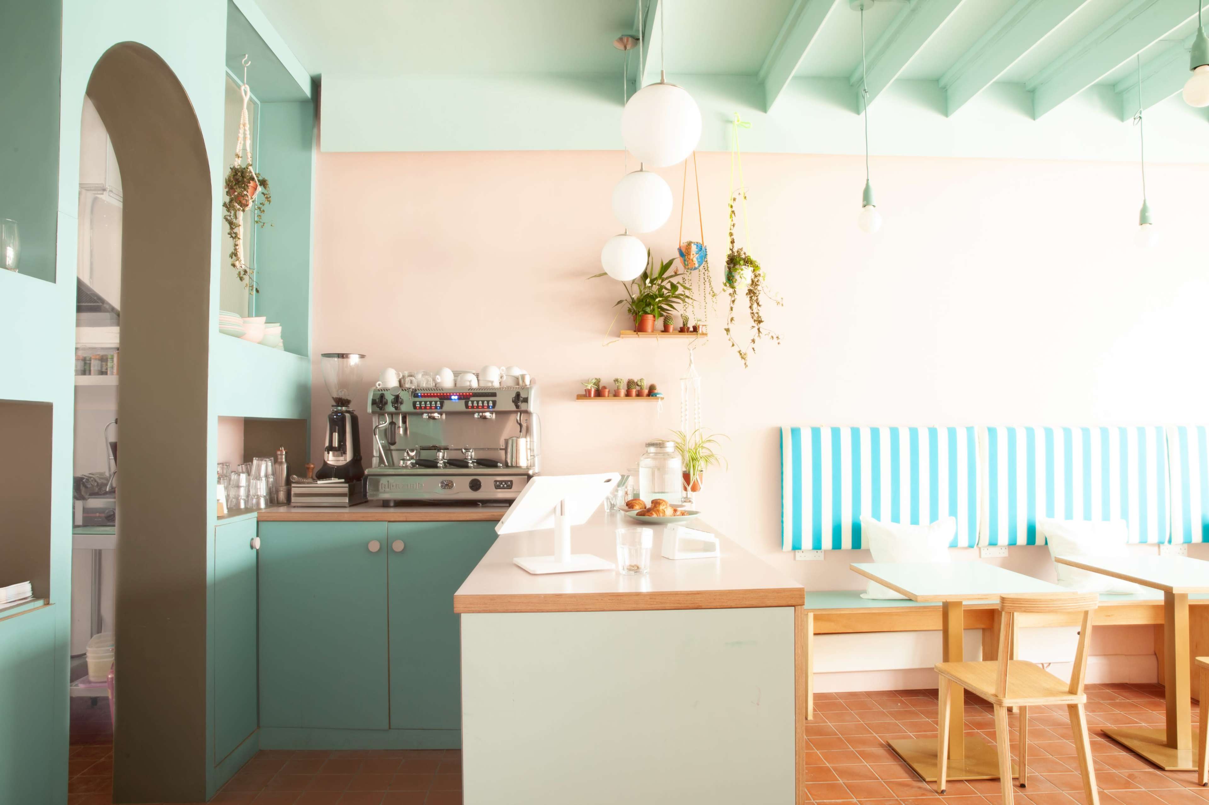 A brightly lit café features teal walls, a coffee machine, and wooden tables with striped seating.