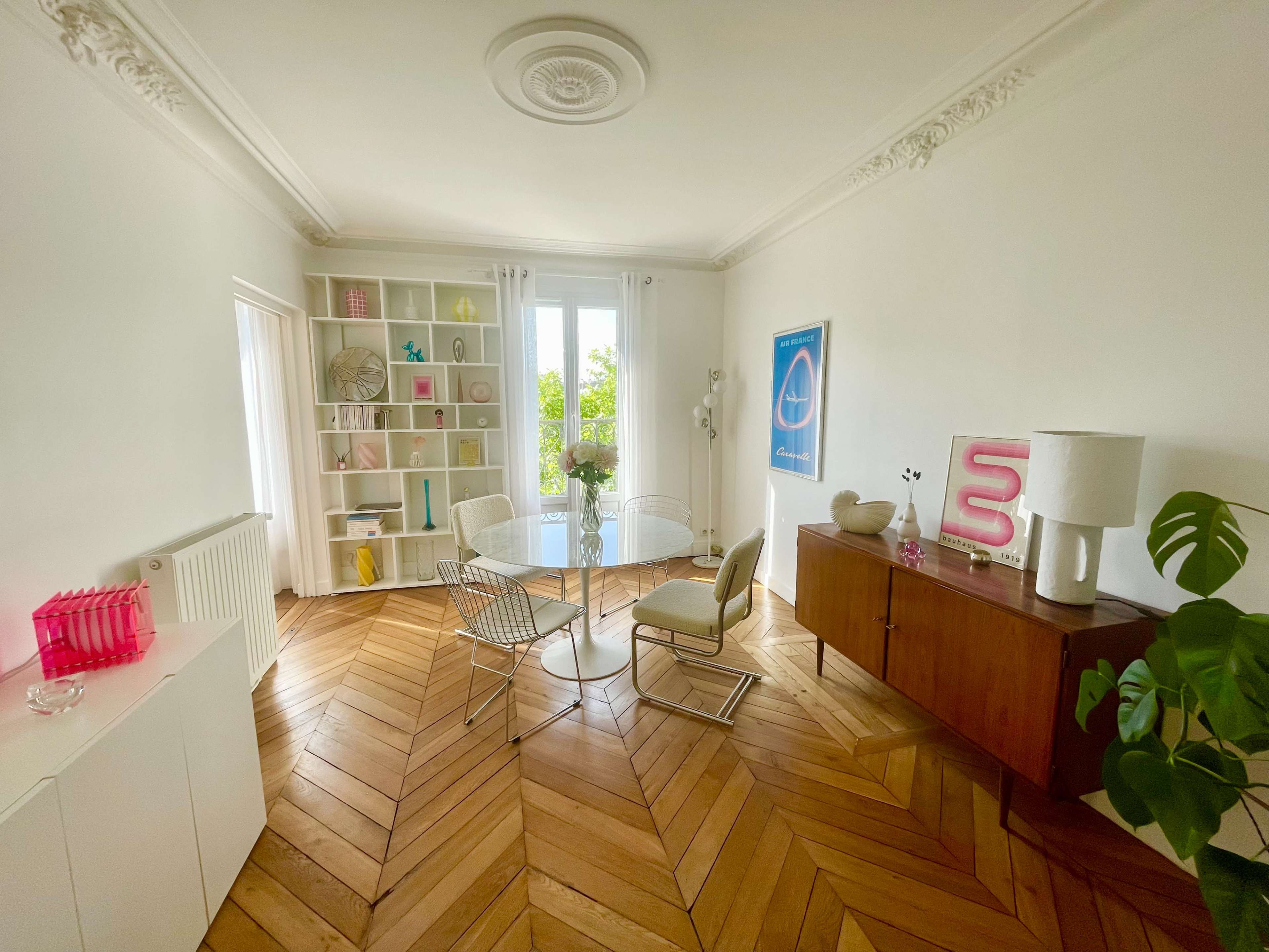 A spacious room features a wooden herringbone floor, a minimalist shelf with decorative items, and a small round table surrounded by two chairs near a window.