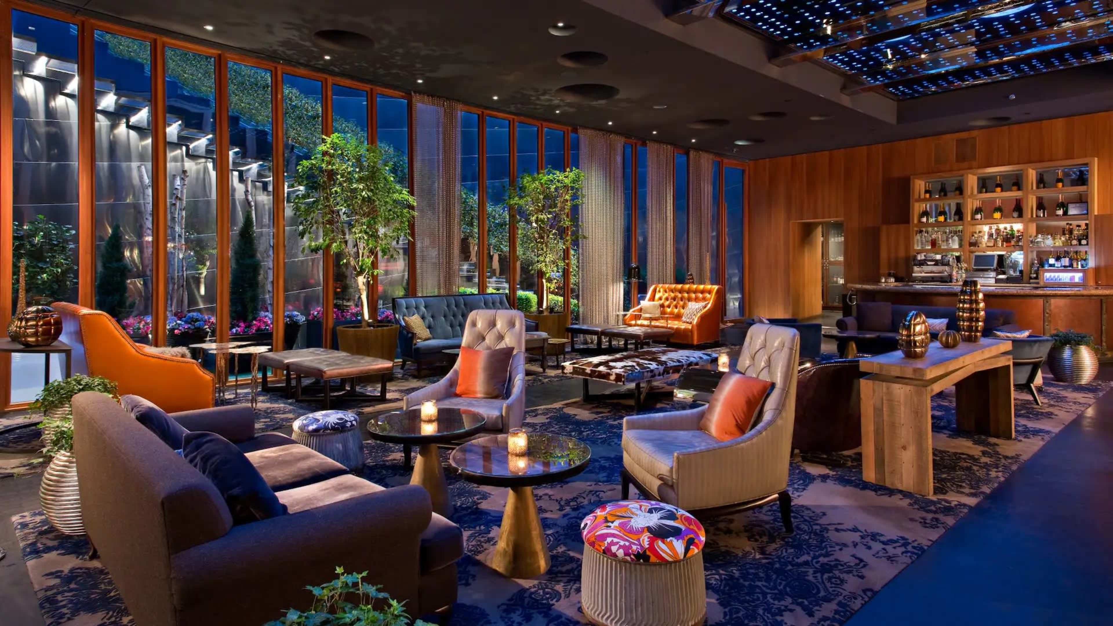 The image shows a modern lounge area with plush seating, decorative plants, and a bar set against large glass windows.