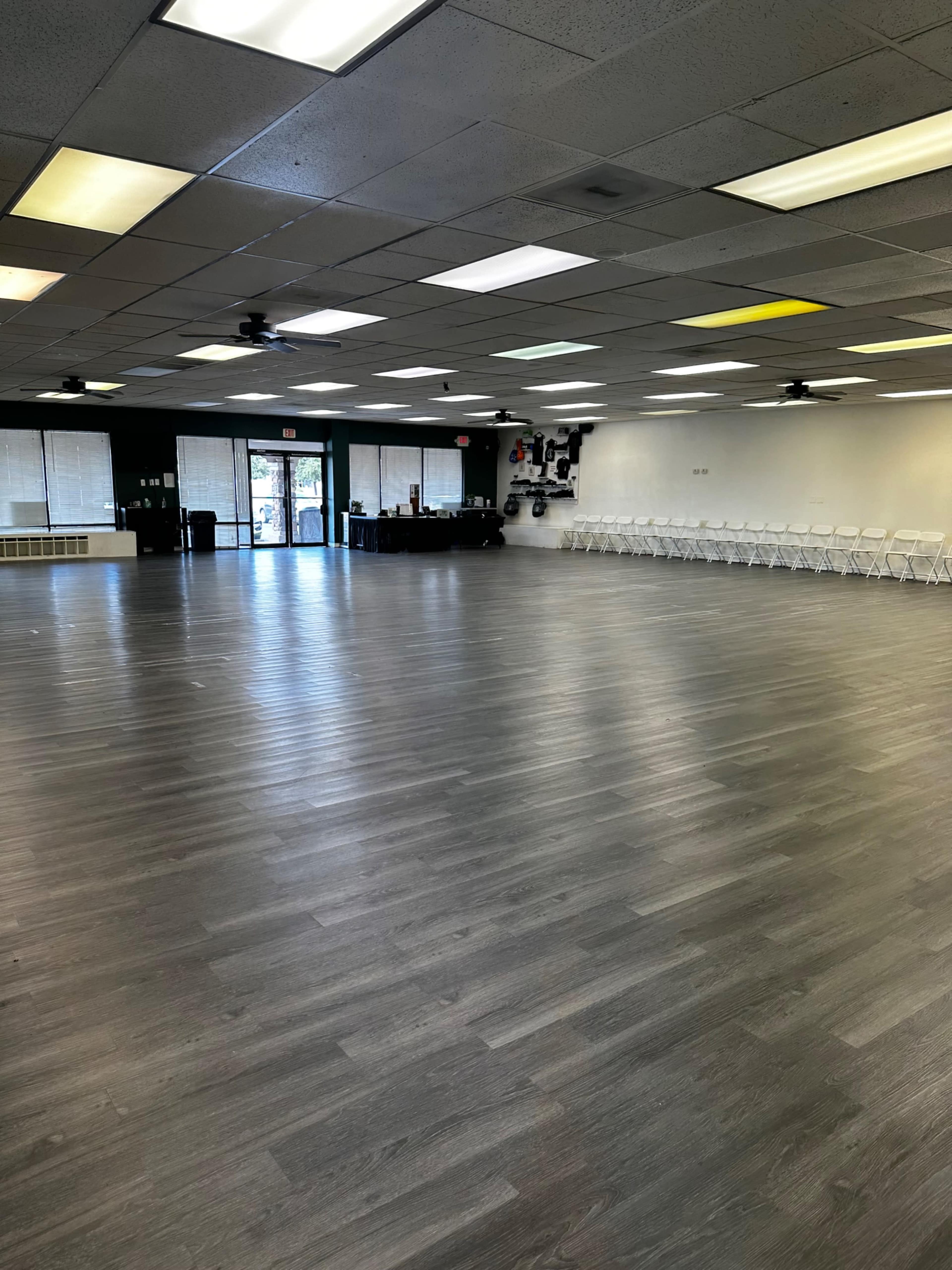 A spacious interior room with a wooden floor, empty chairs lined against one wall, and large windows providing natural light.