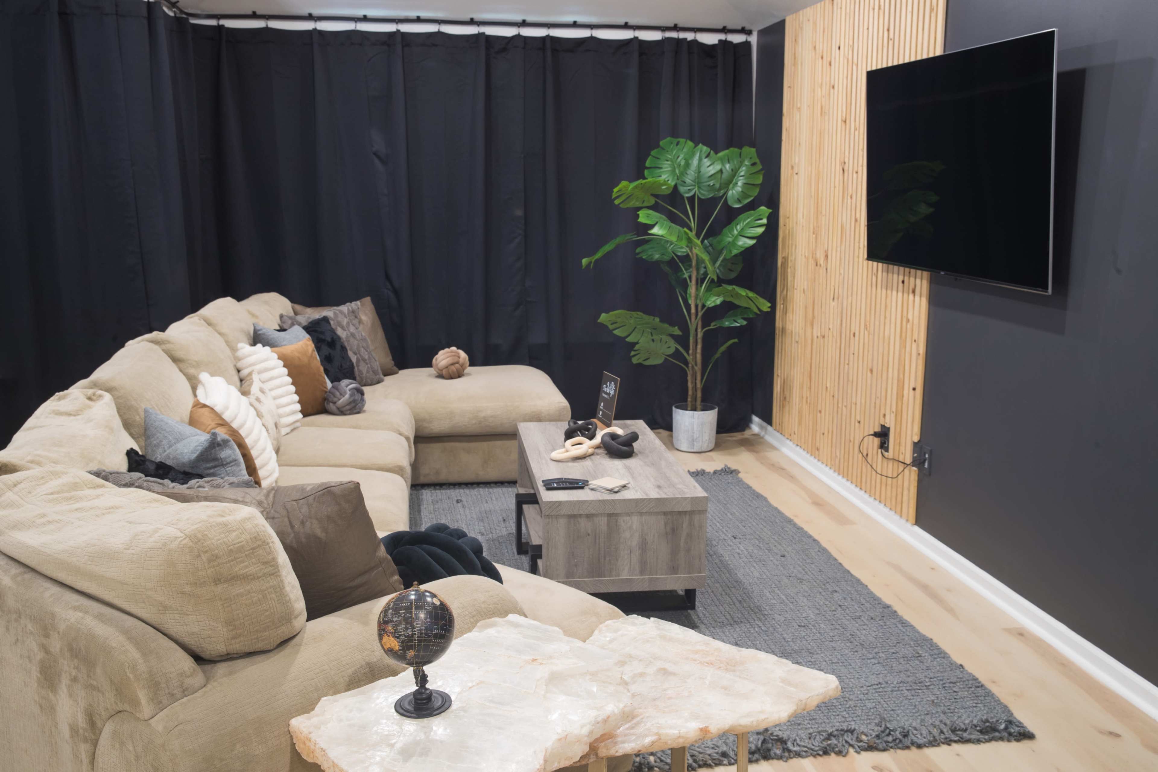 A modern living room features a large sectional sofa, a coffee table, a potted plant, and a wall-mounted television against a dark backdrop.
