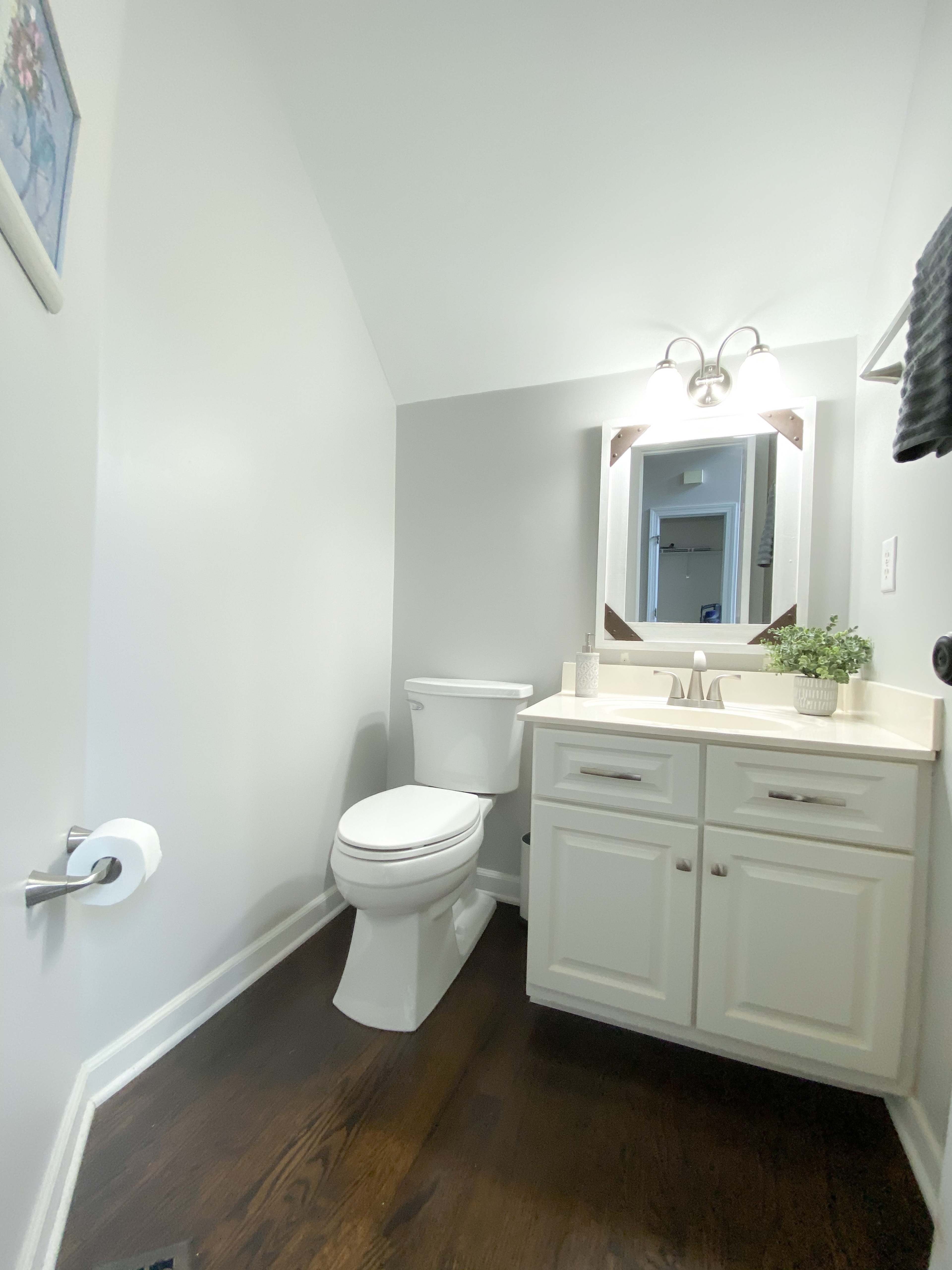 A small bathroom with a white toilet, a light-colored vanity with a sink, and a mirror above it, all set against gray walls and dark wood flooring.