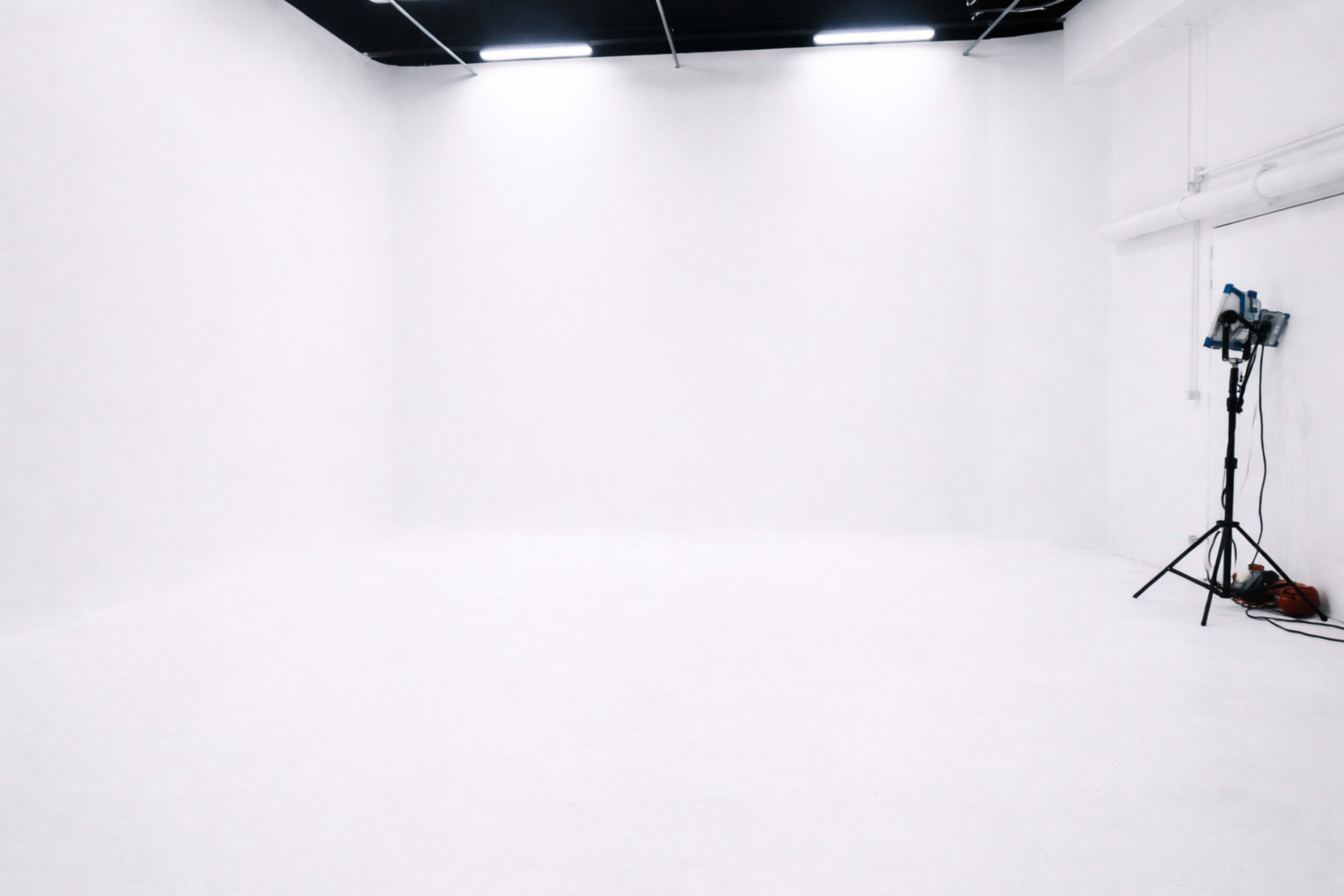 The image shows a large, empty white room with smooth walls and a tripod lighting setup in one corner.