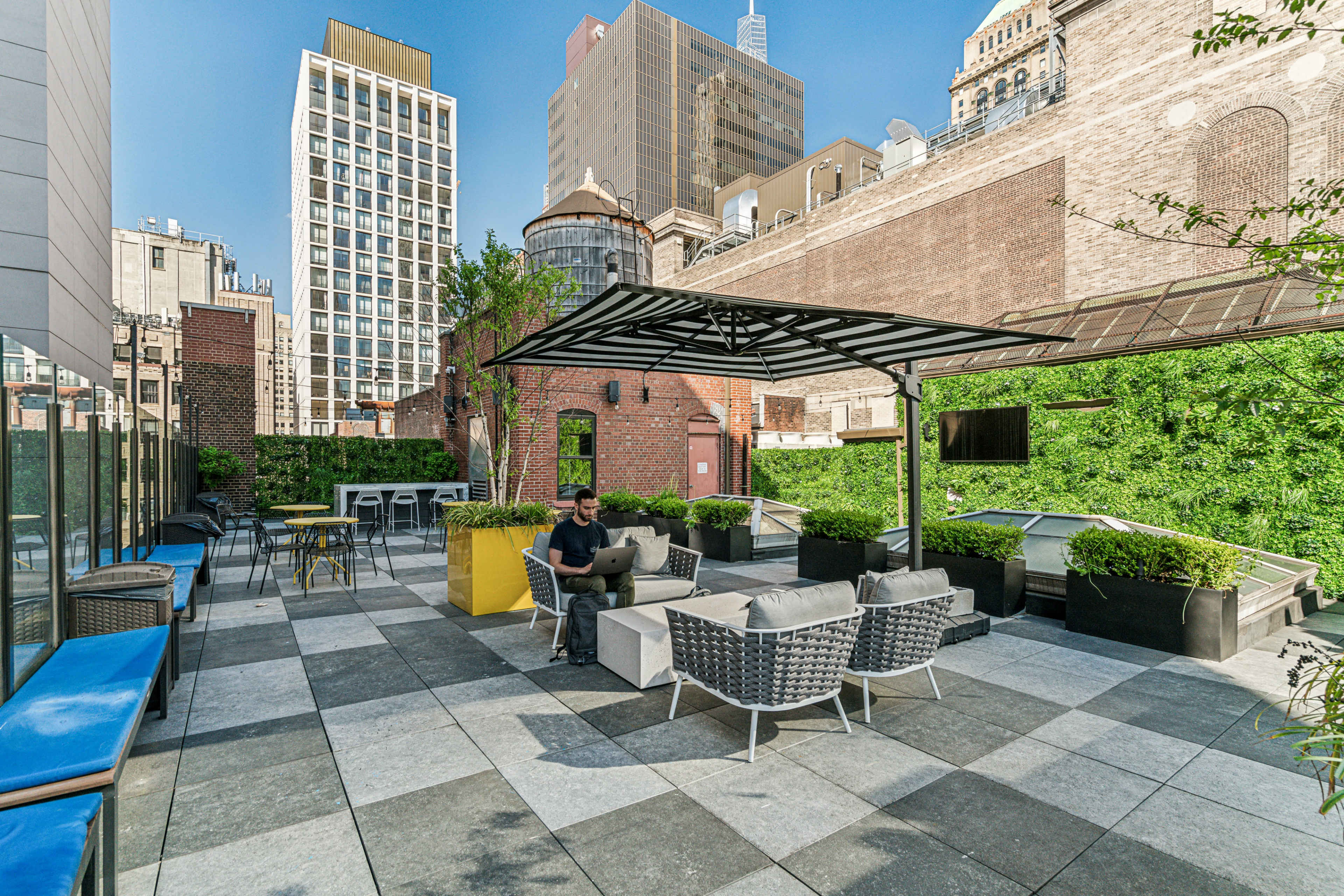 A person is seated on a patio with modern furniture and greenery, surrounded by high-rise buildings.