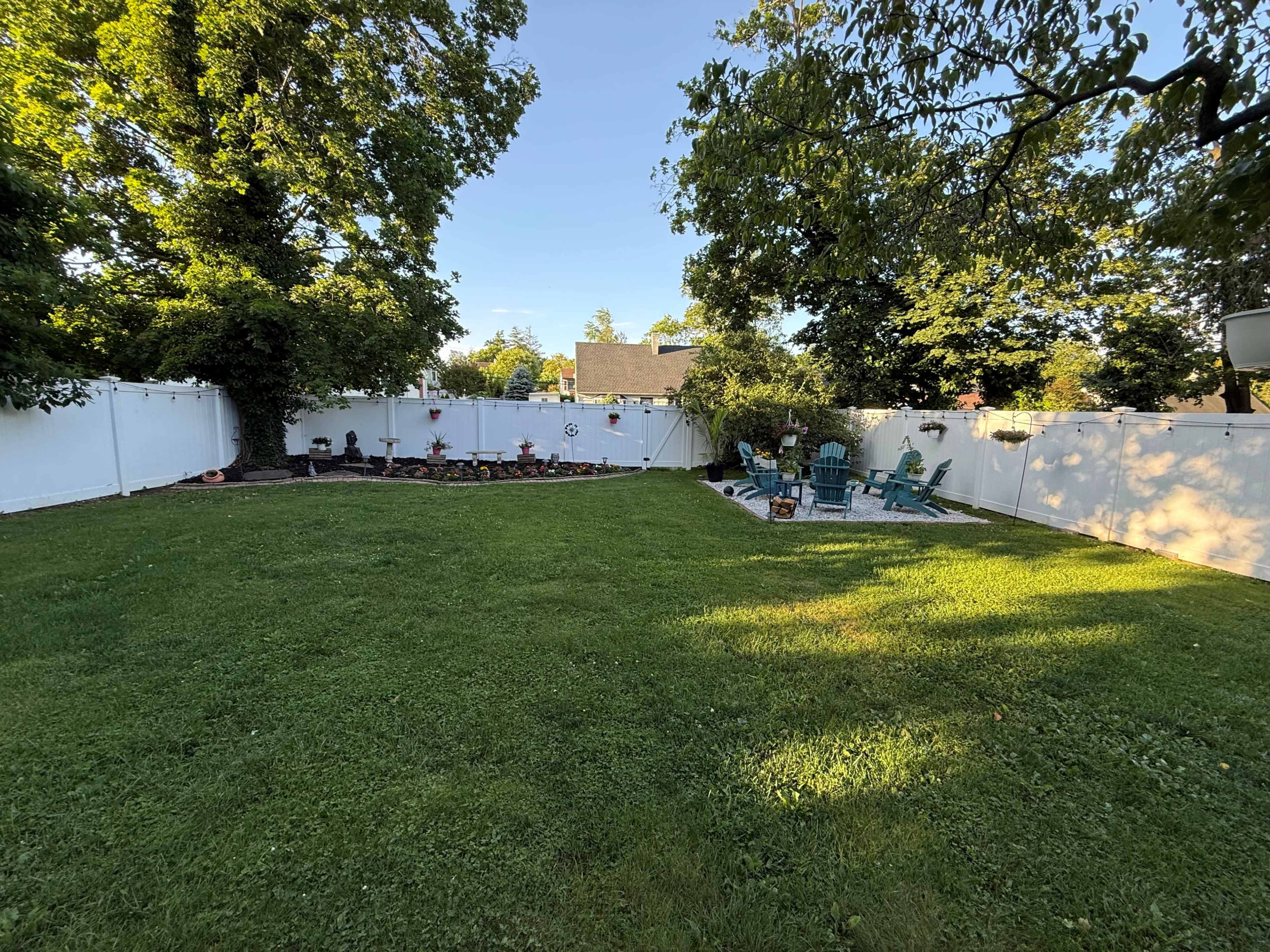The image shows a well-maintained backyard with a grassy lawn, a white fence, and seating arrangements under trees.