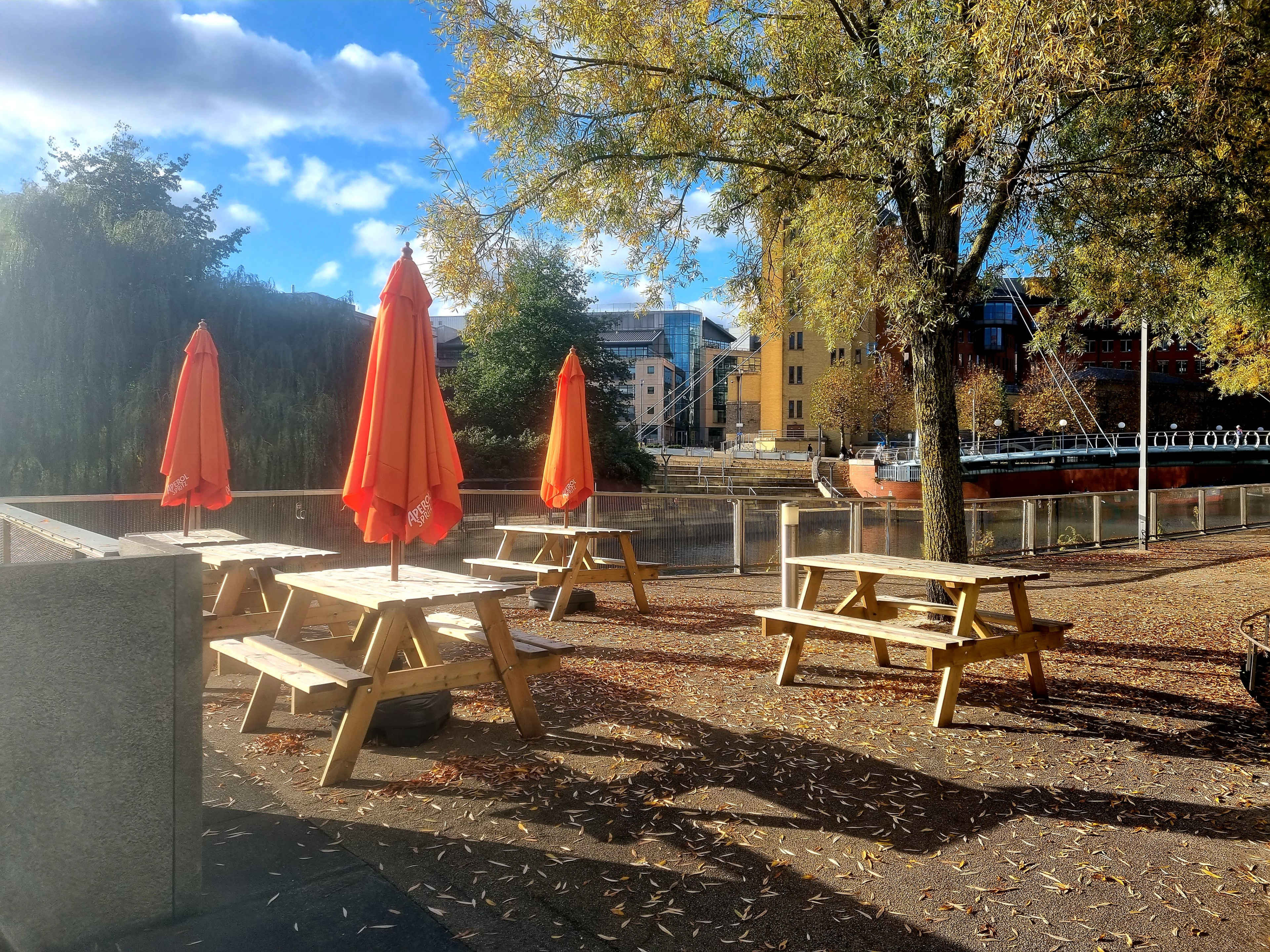 The scene features several wooden picnic tables under bright orange umbrellas near a calm river surrounded by trees and buildings.