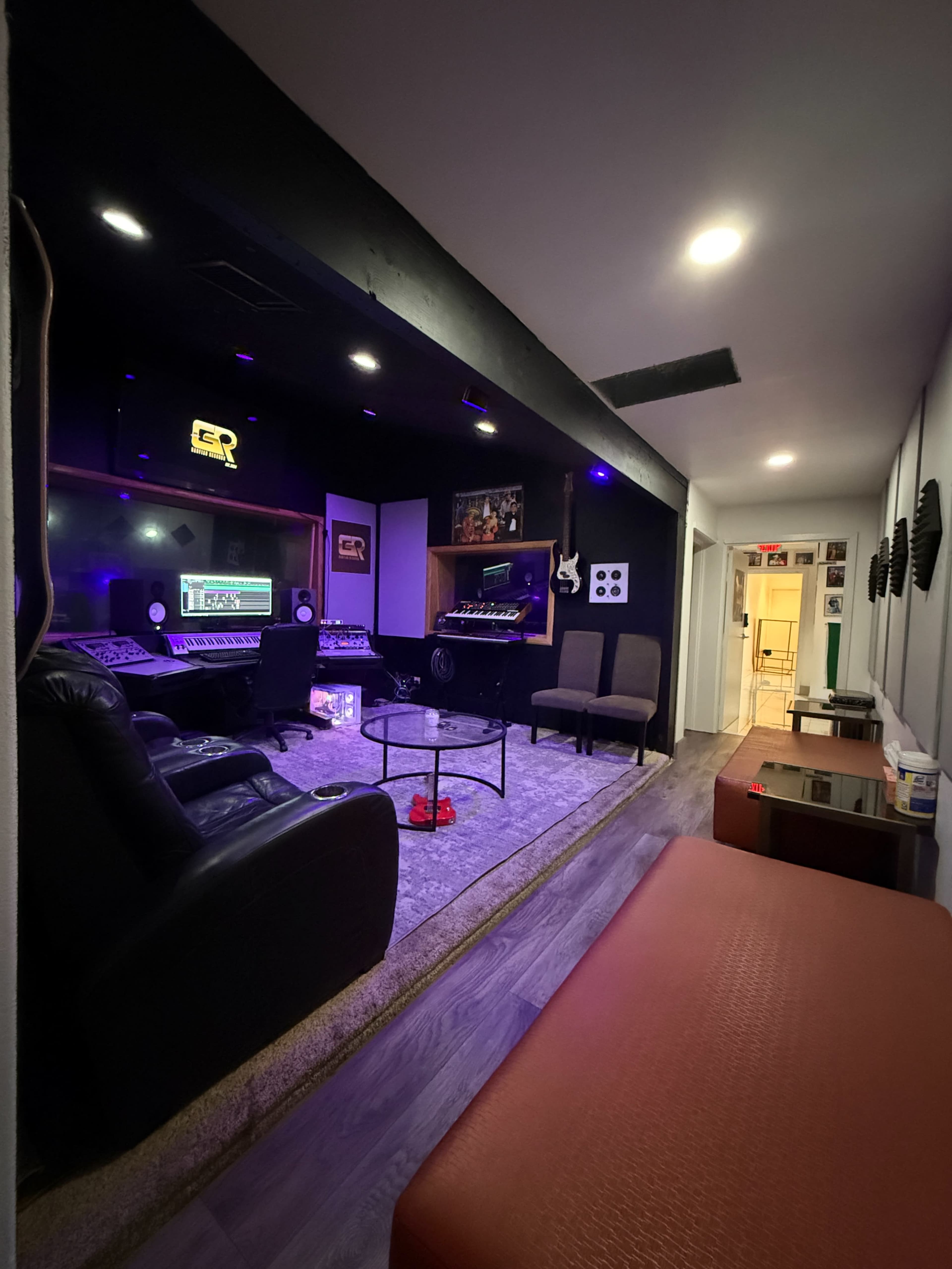 The image shows a recording studio with a control desk, comfortable seating, and soundproofing panels on the walls.