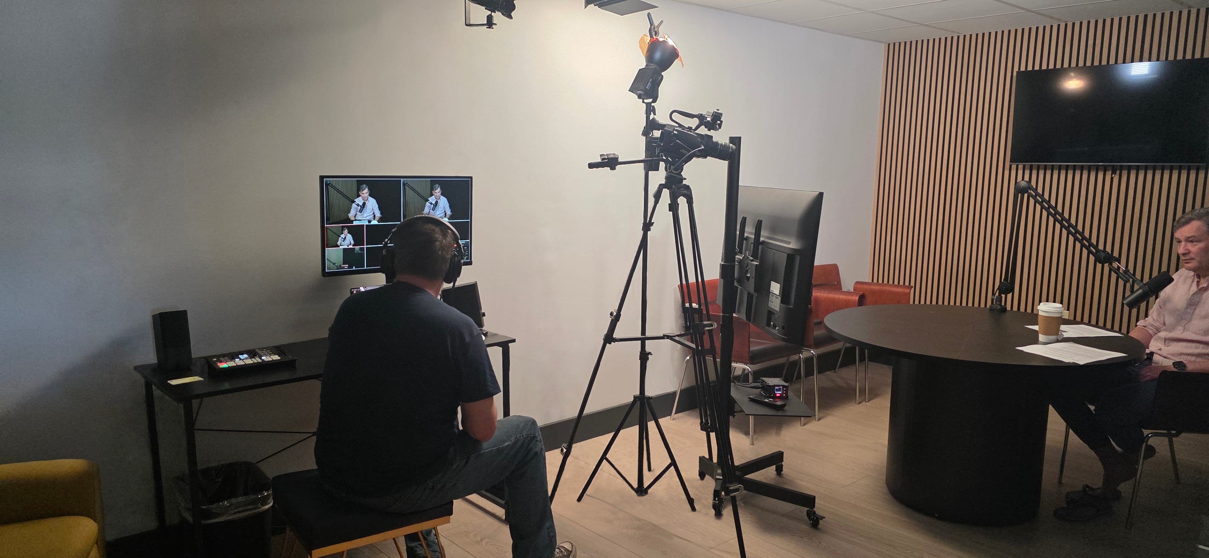 A person sits at a control station monitoring a video production setup featuring multiple screens and cameras in a modern studio space.