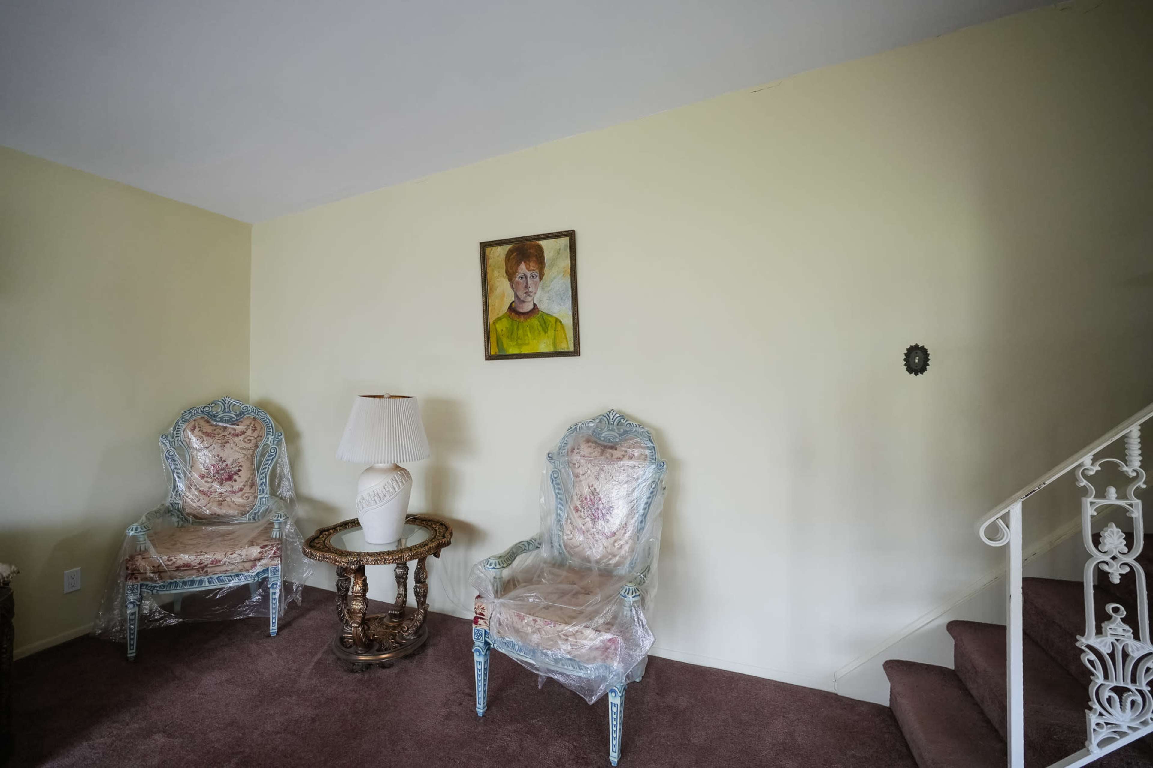 The image shows a small room with two ornate chairs and a round table with a lamp, along with a painting on the wall and a staircase leading up.