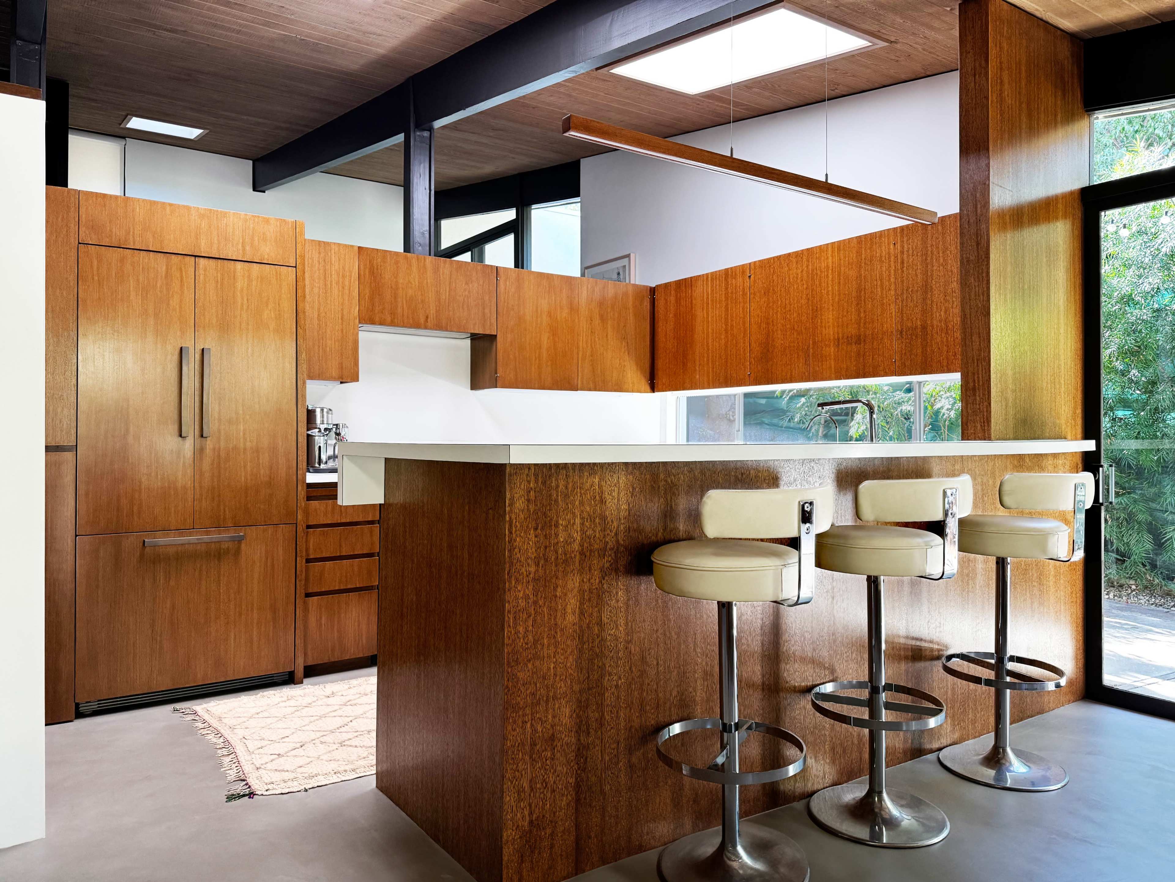 The image shows a modern kitchen featuring wooden cabinets, a central island with three bar stools, and large windows that allow natural light to enter.