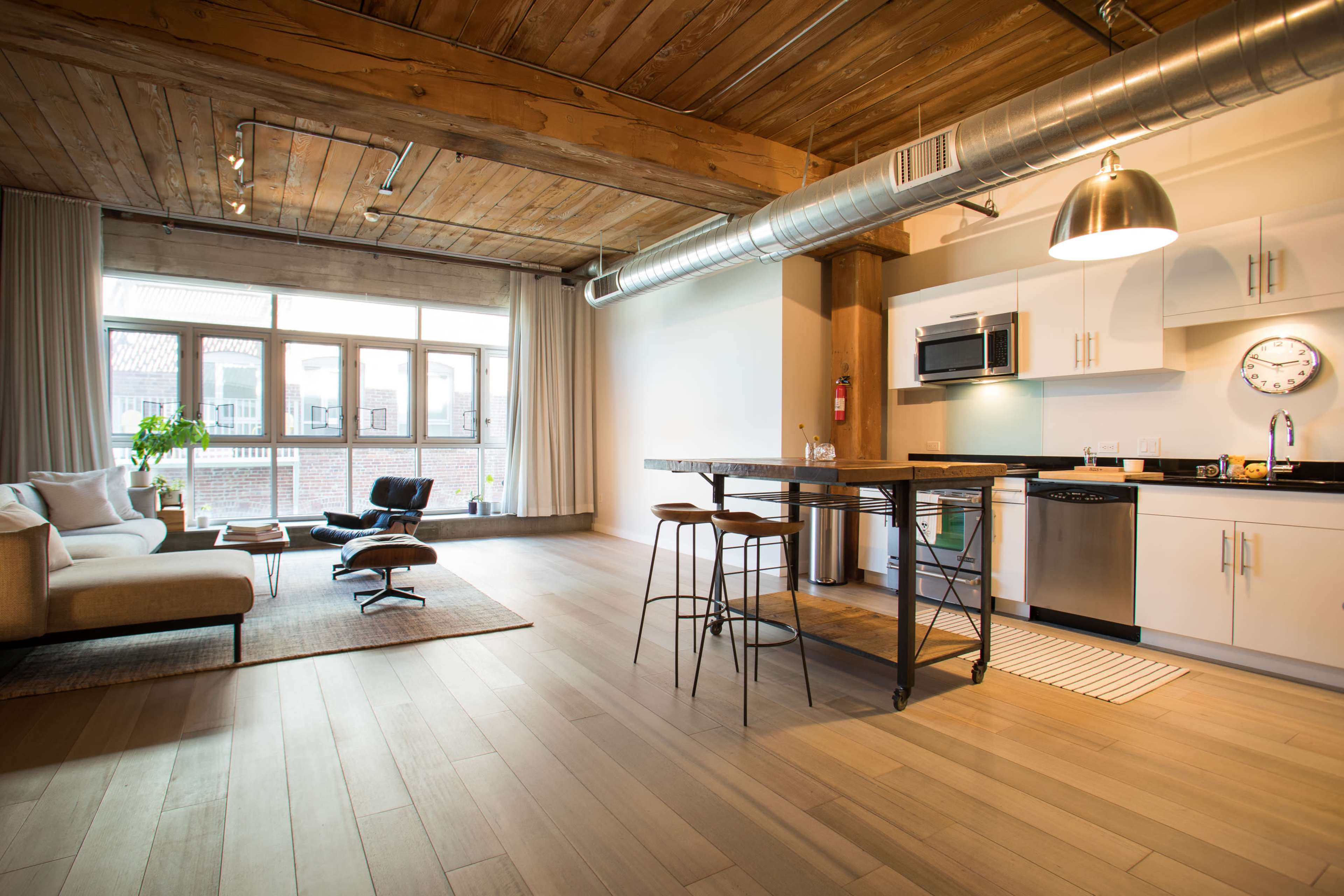 The image shows a modern apartment interior featuring a living area with a couch, a kitchen with stainless steel appliances, and large windows allowing natural light to fill the space.
