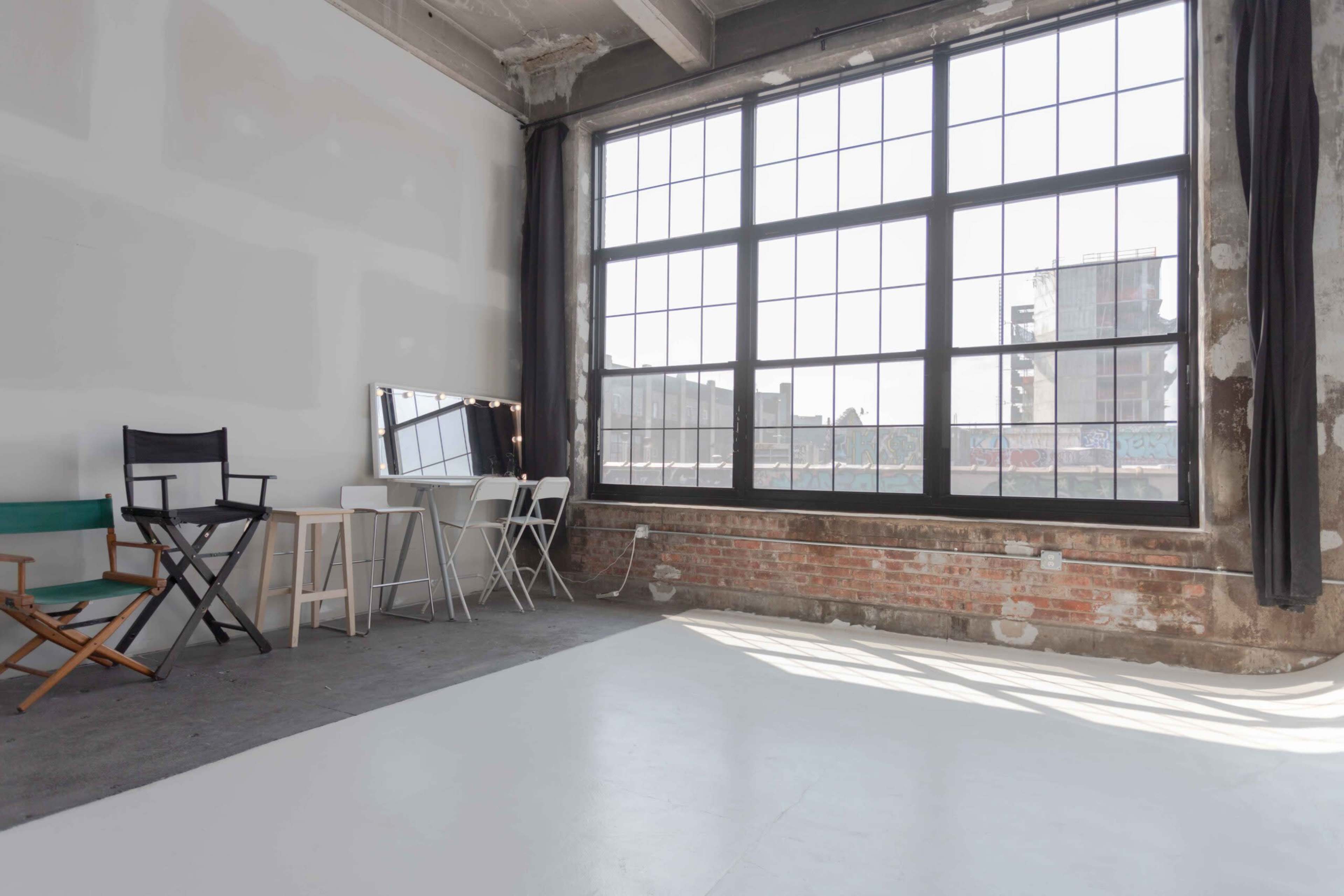 The image shows a spacious, well-lit interior with large windows, brick walls, and a few minimalist pieces of furniture including chairs and a mirror.