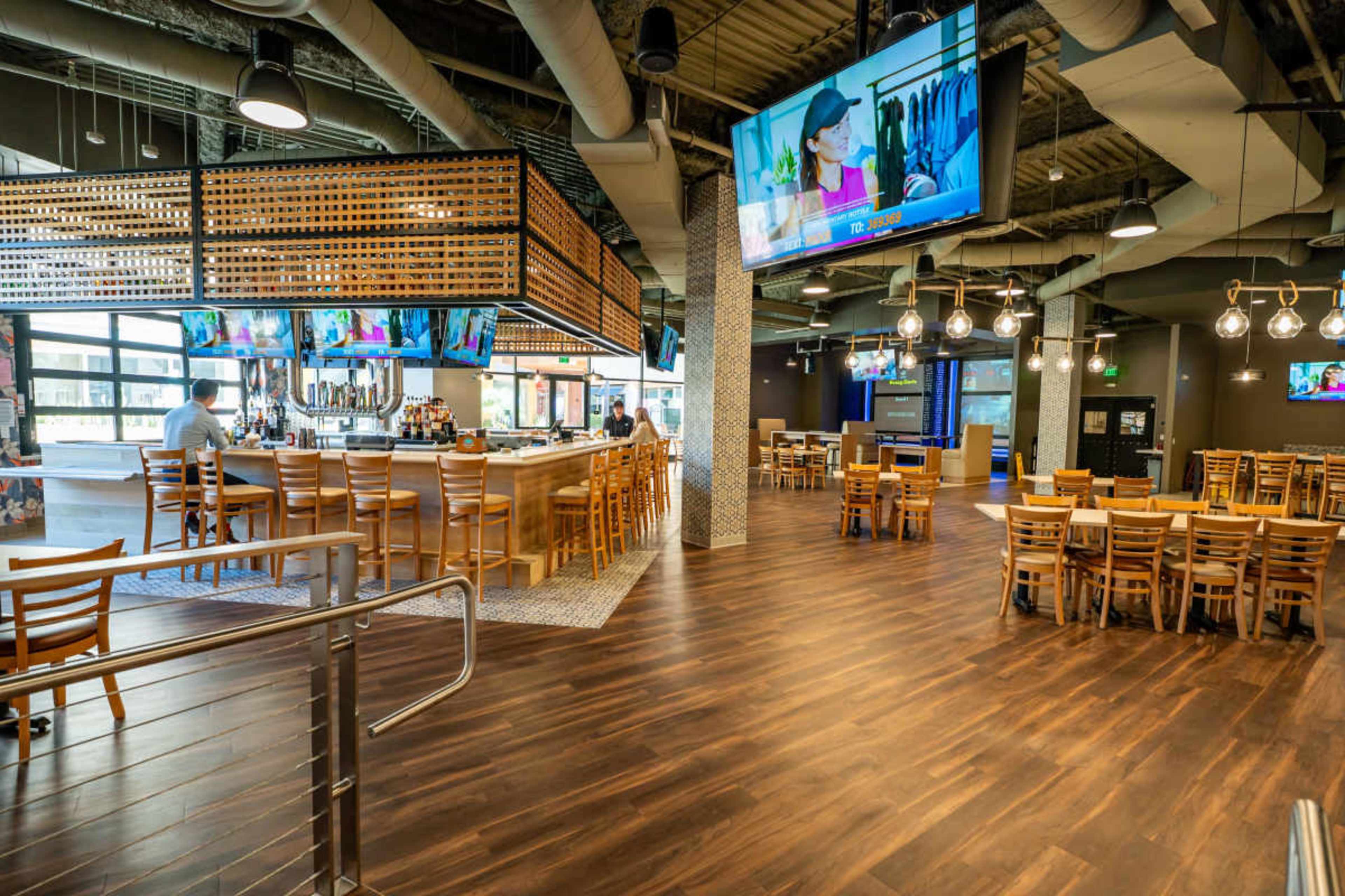 The image shows the interior of a modern restaurant with a bar, multiple televisions, and wooden tables and chairs arranged for dining.