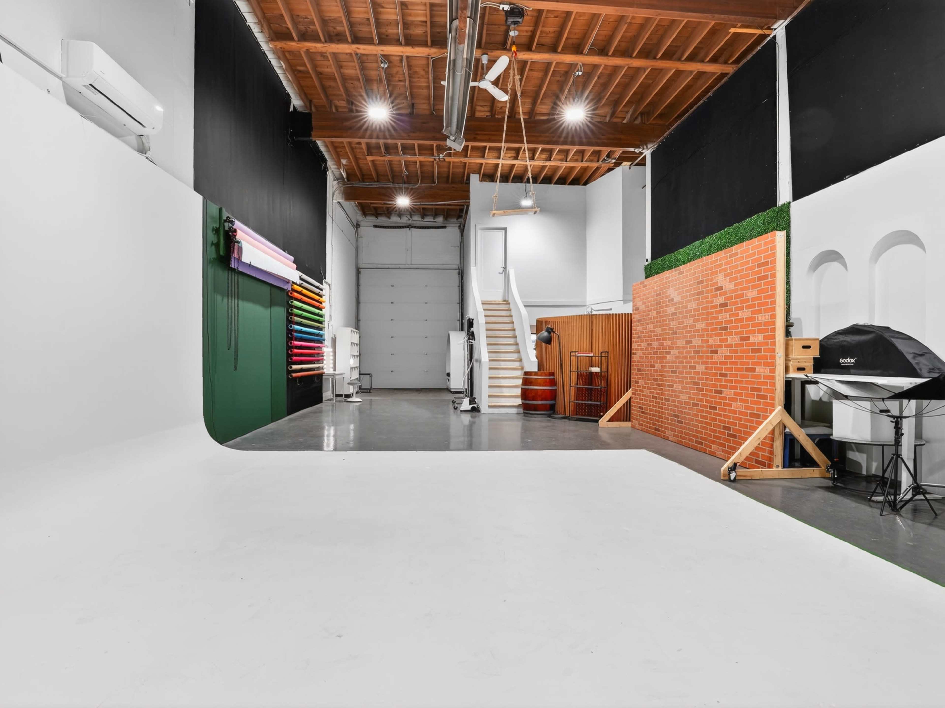 The image shows a spacious photography studio with a white backdrop, colorful roll paper backgrounds, a staircase, and various props against the walls.