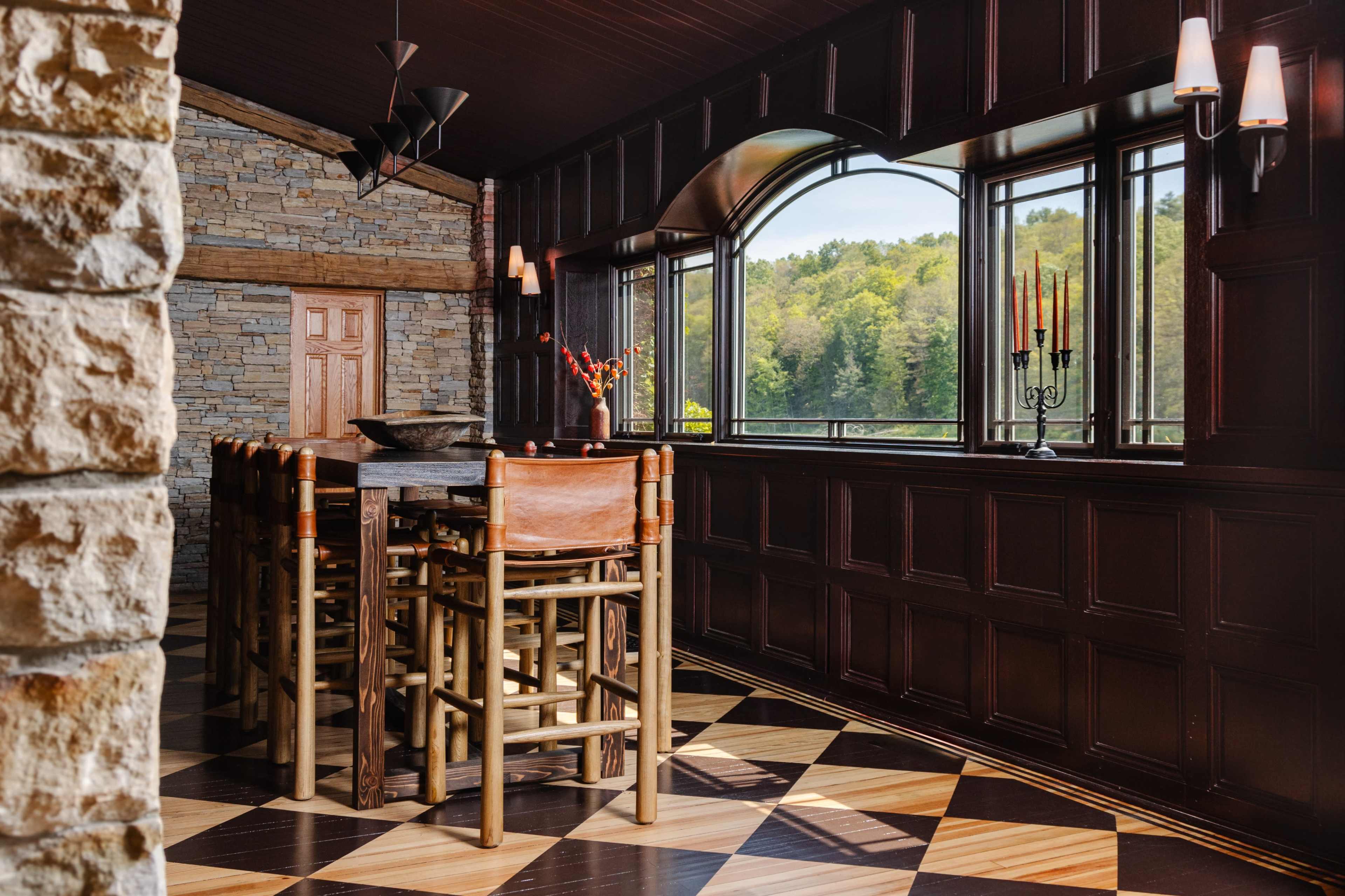 A modern dining area with a large wooden table surrounded by tall chairs, large windows overlooking a natural landscape, and decorative candle holders on the walls.