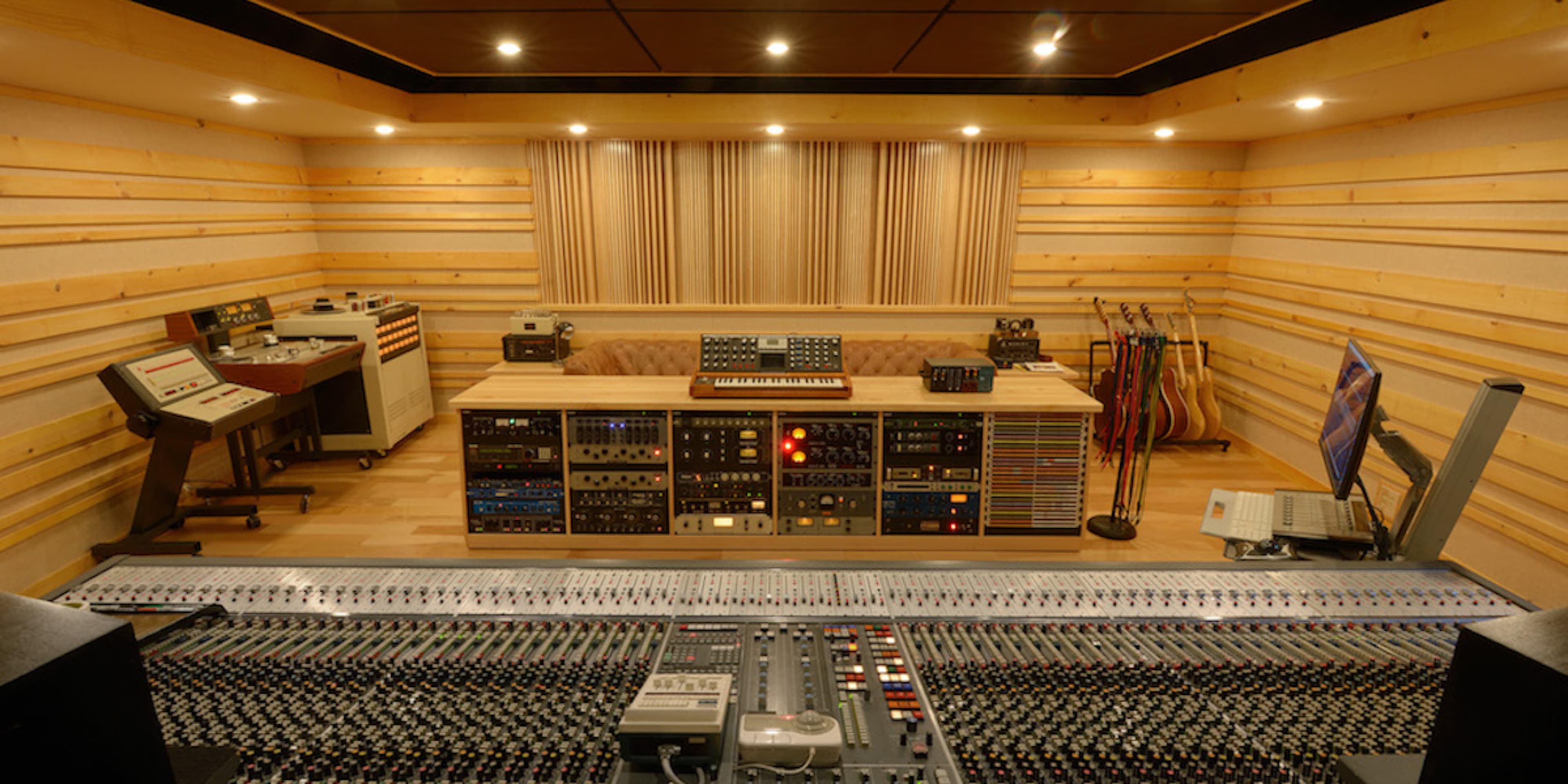 A recording studio with a large mixing console, various audio equipment, and instruments, set against wooden paneling.
