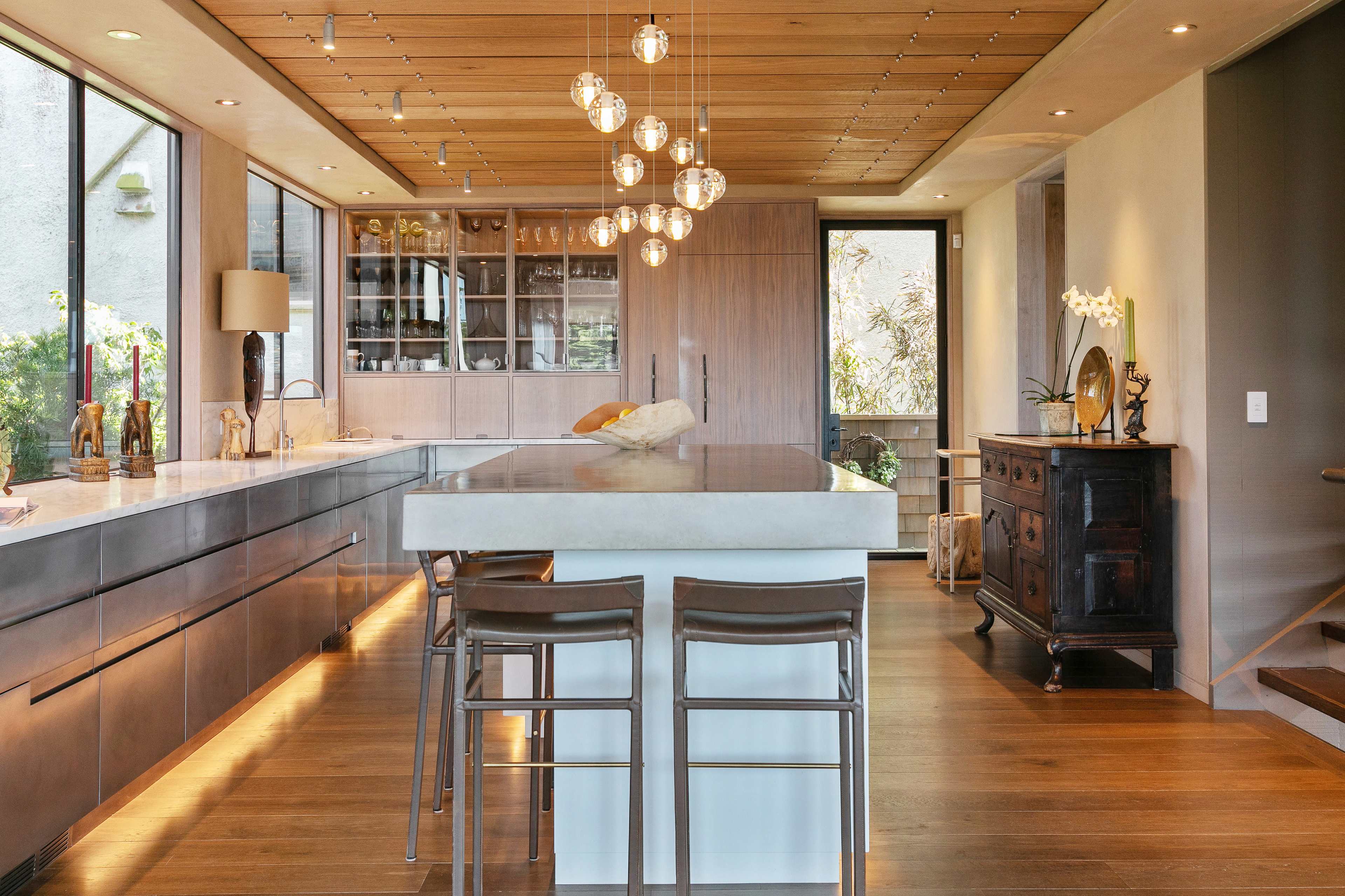 A modern kitchen features a long island with seating, pendant lighting, and large windows showing greenery outside.