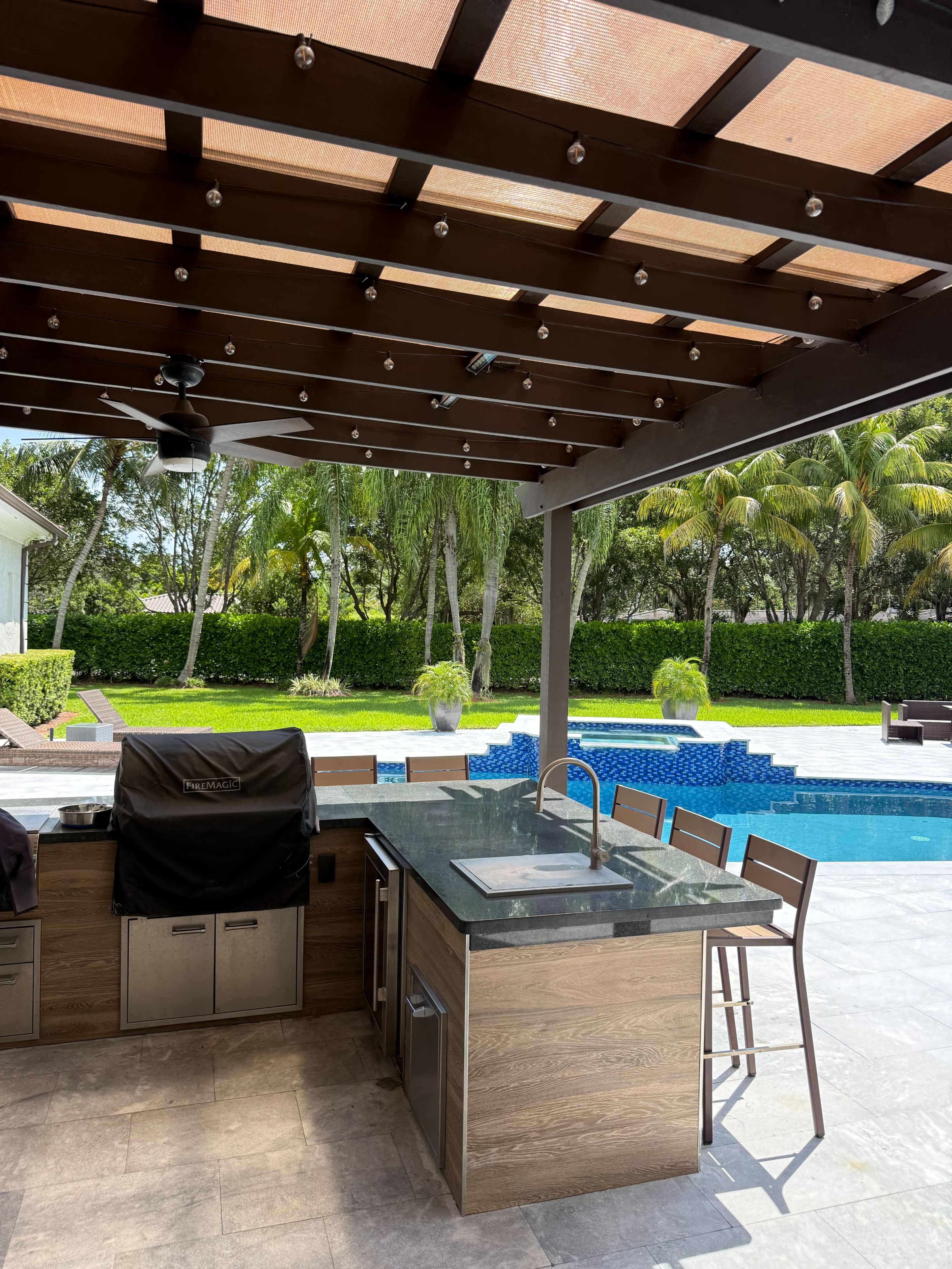 The image shows a modern outdoor kitchen and seating area under a wooden pergola beside a swimming pool surrounded by palm trees and landscaping.