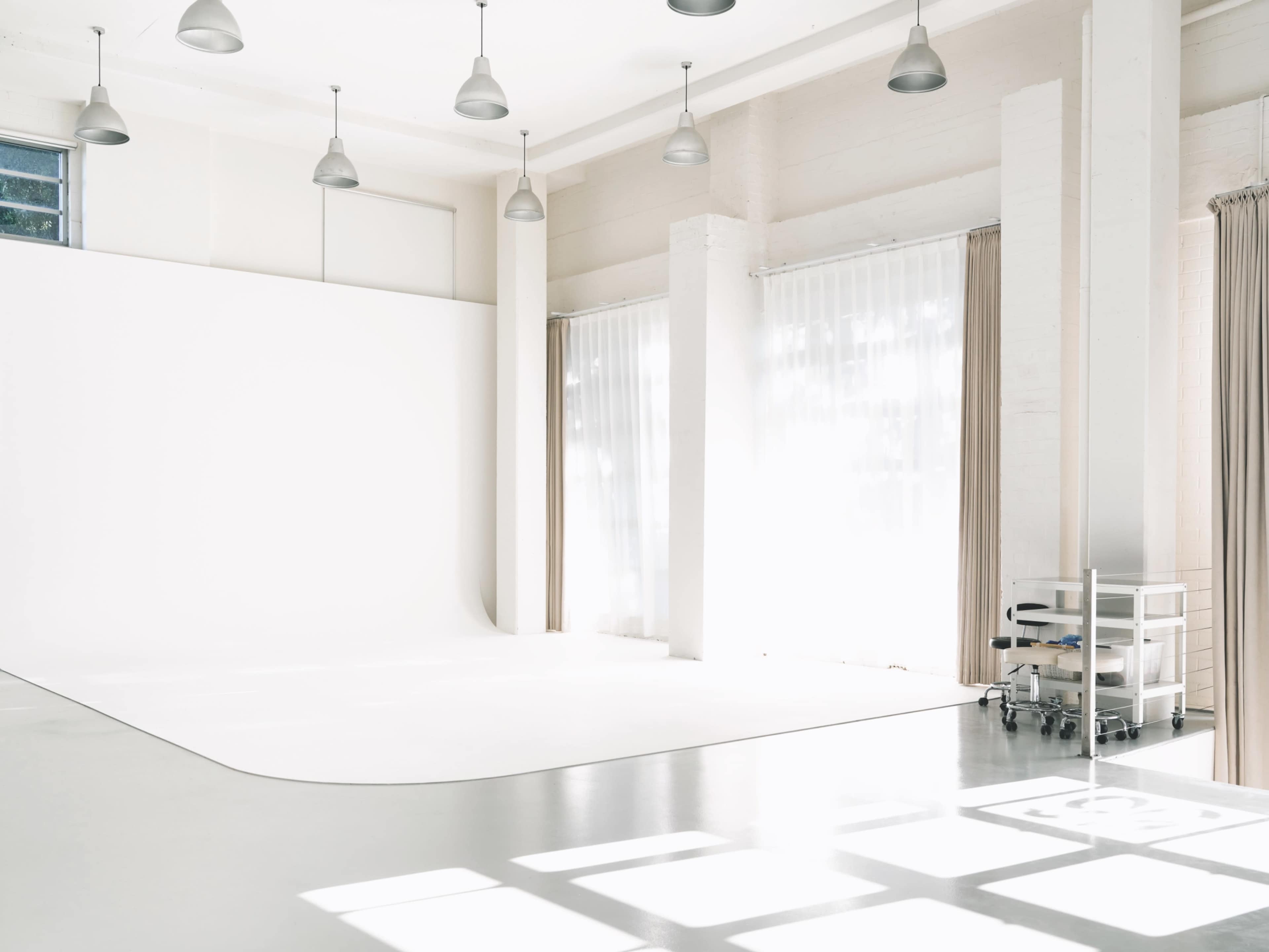 The image shows a bright, minimalist studio with a large white backdrop, large windows letting in natural light, and light-colored curtains.