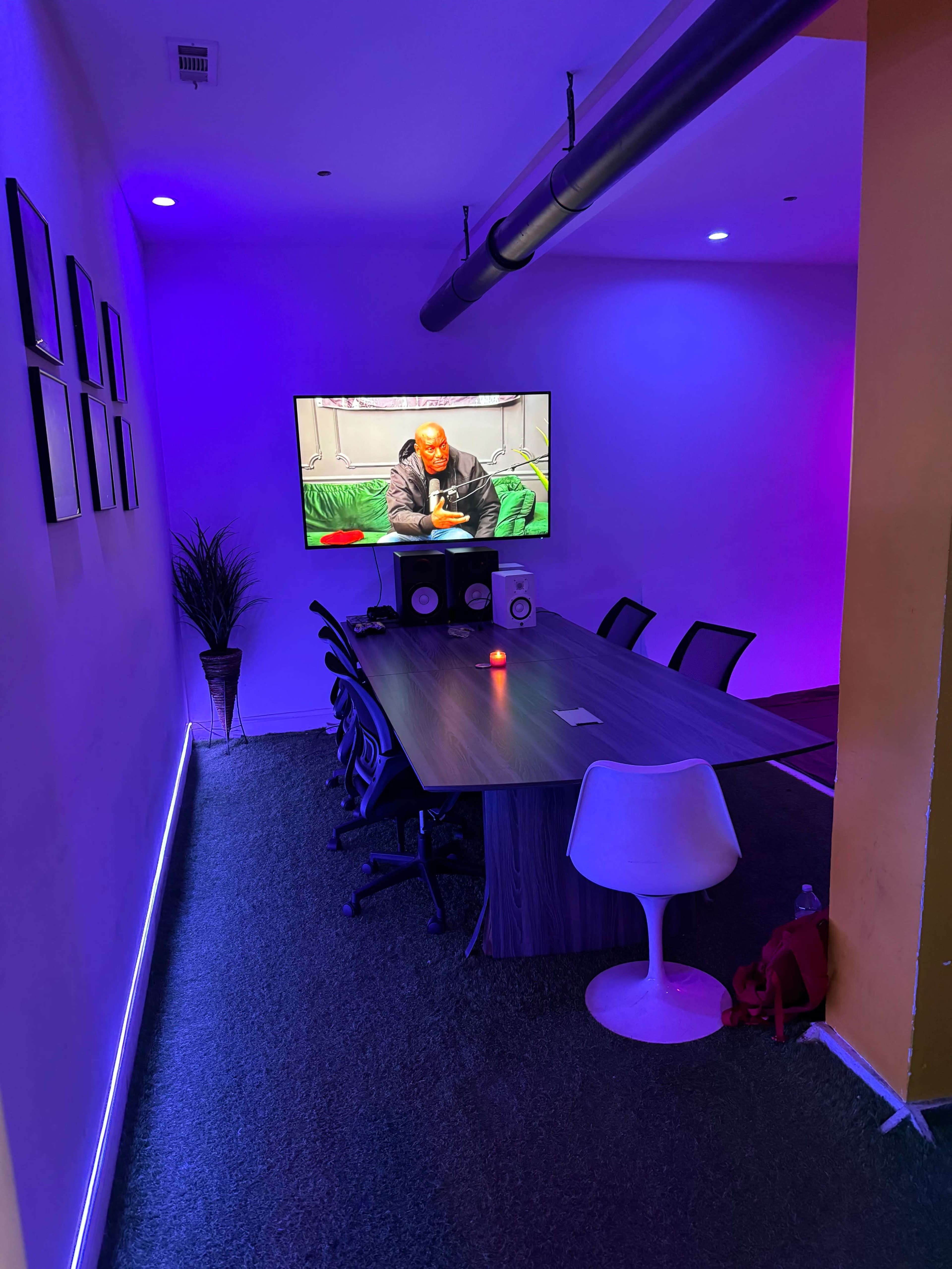 A modern meeting room features a long wooden table, black chairs, a large wall-mounted TV displaying a scene, and colorful ambient lighting.