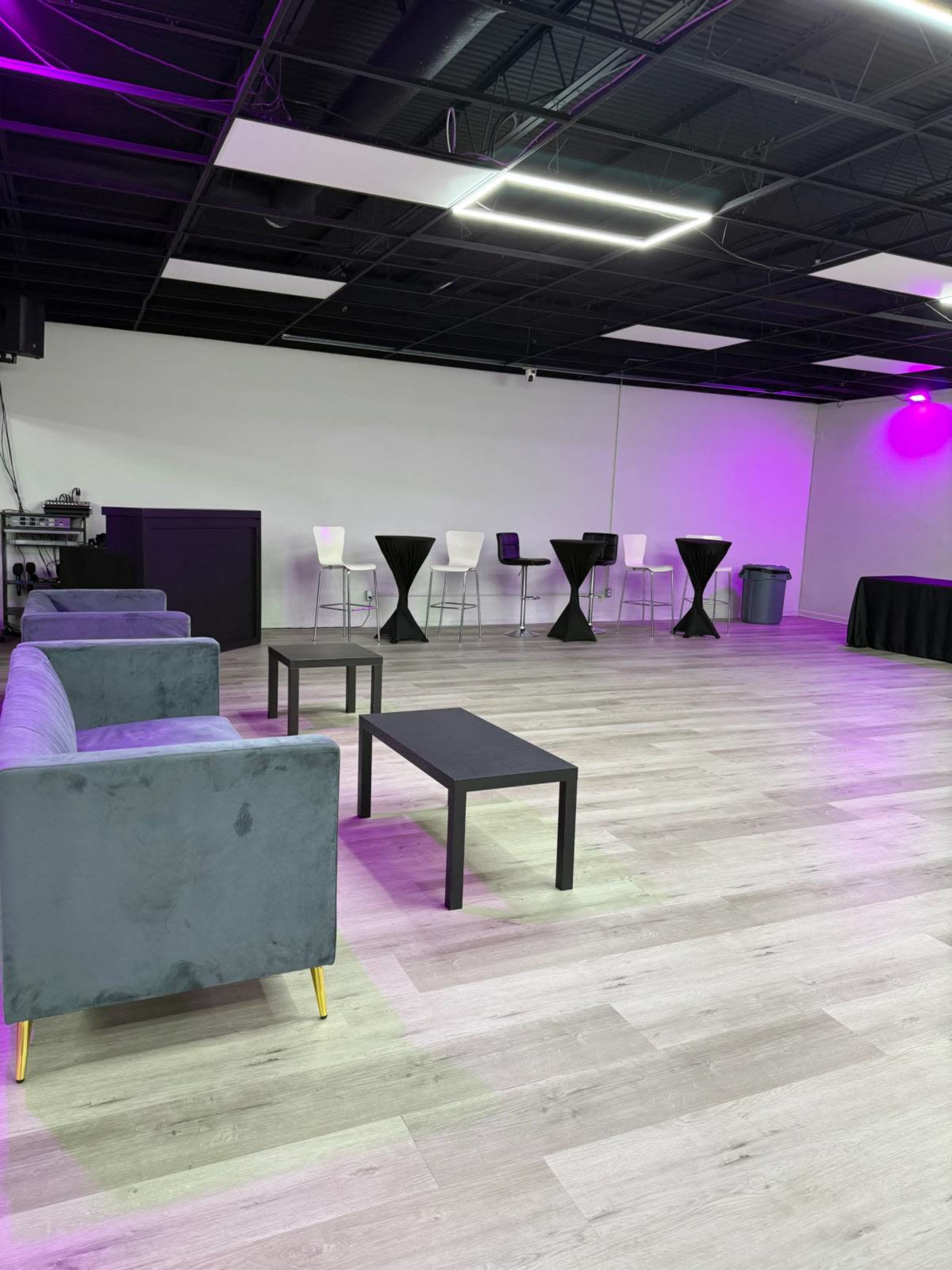 The image shows a spacious room with grey couches, black tables, and a bar area illuminated by purple lights.