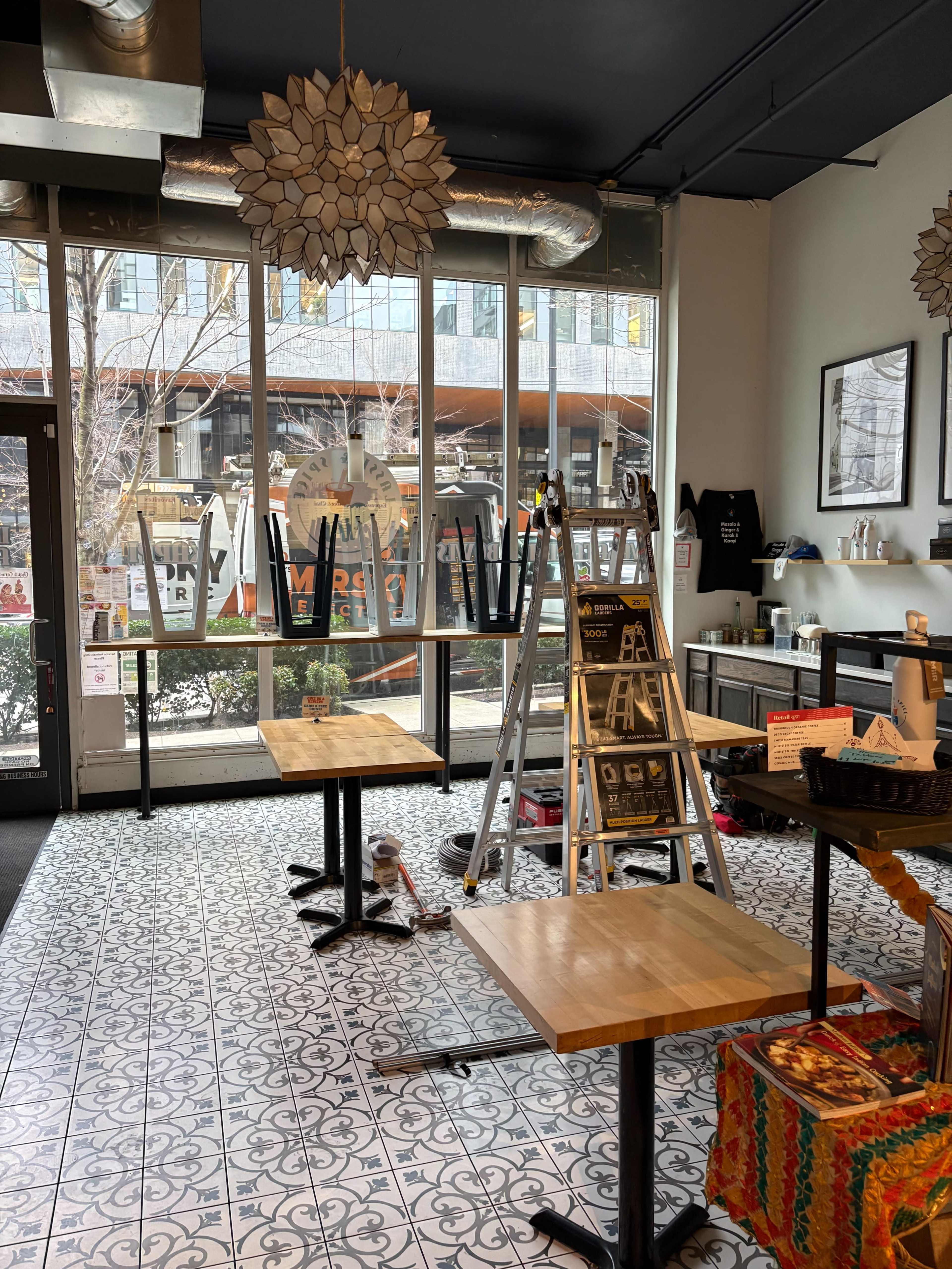 The interior of a cafe features two wooden tables, a ladder, and large windows displaying a city view.