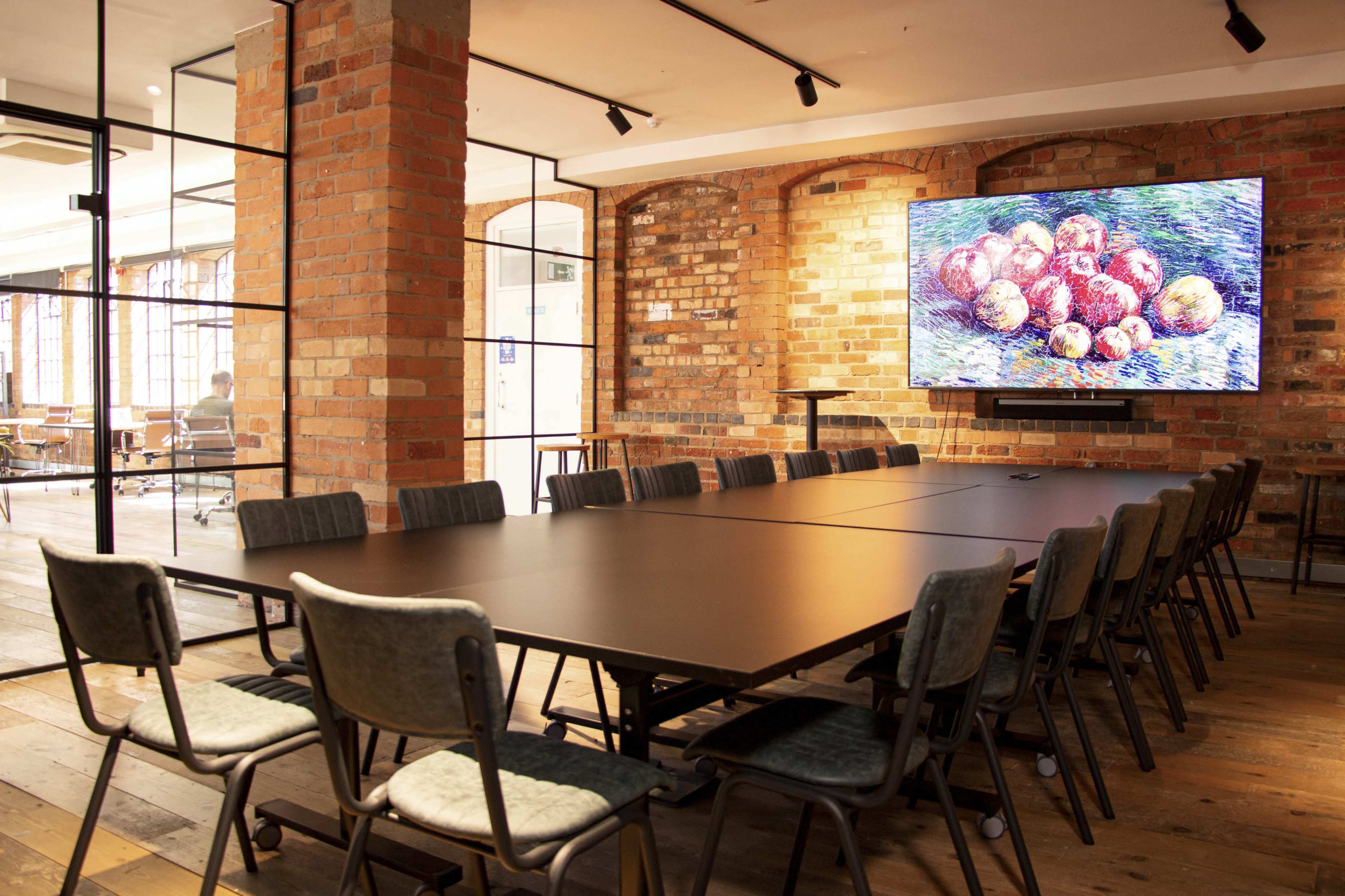 A modern conference room features a large table surrounded by chairs, with an artwork displayed on a wall and glass partitions in the background.