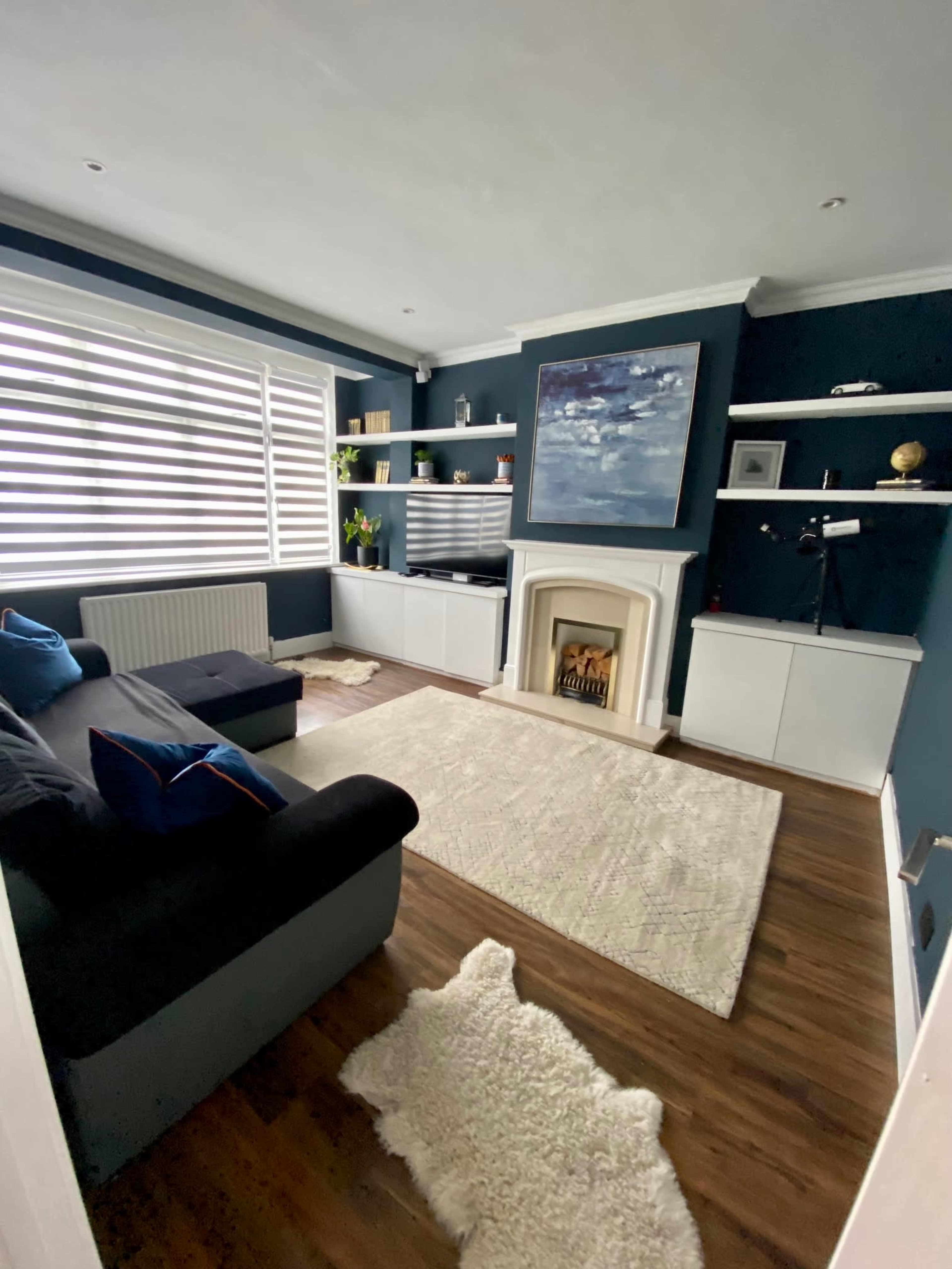 A cozy living room with dark blue walls, modern furniture, a fireplace, and a large window with striped blinds.