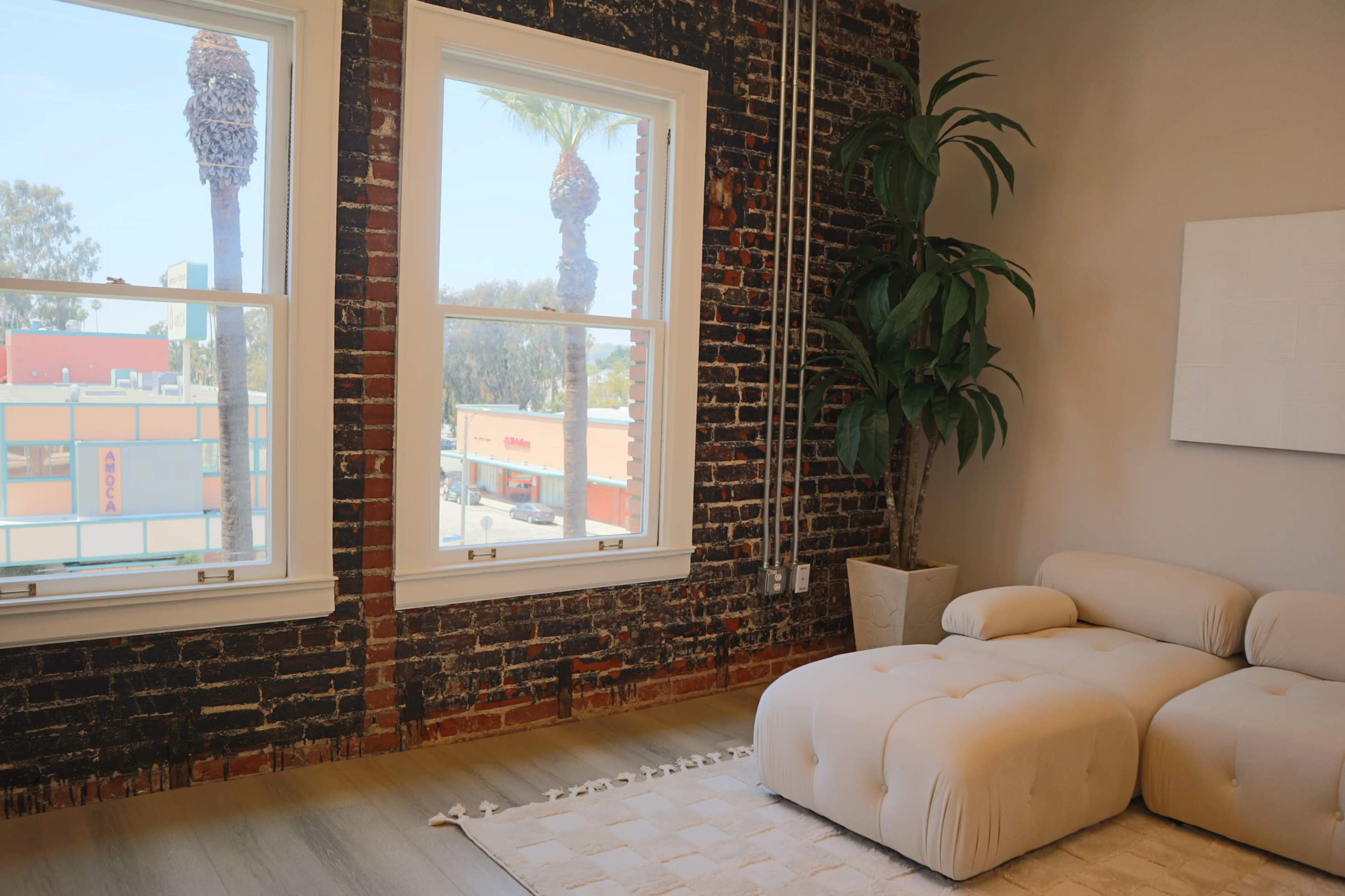 A cozy interior featuring a light-colored sectional sofa, large windows with palm trees visible outside, and exposed brick walls.