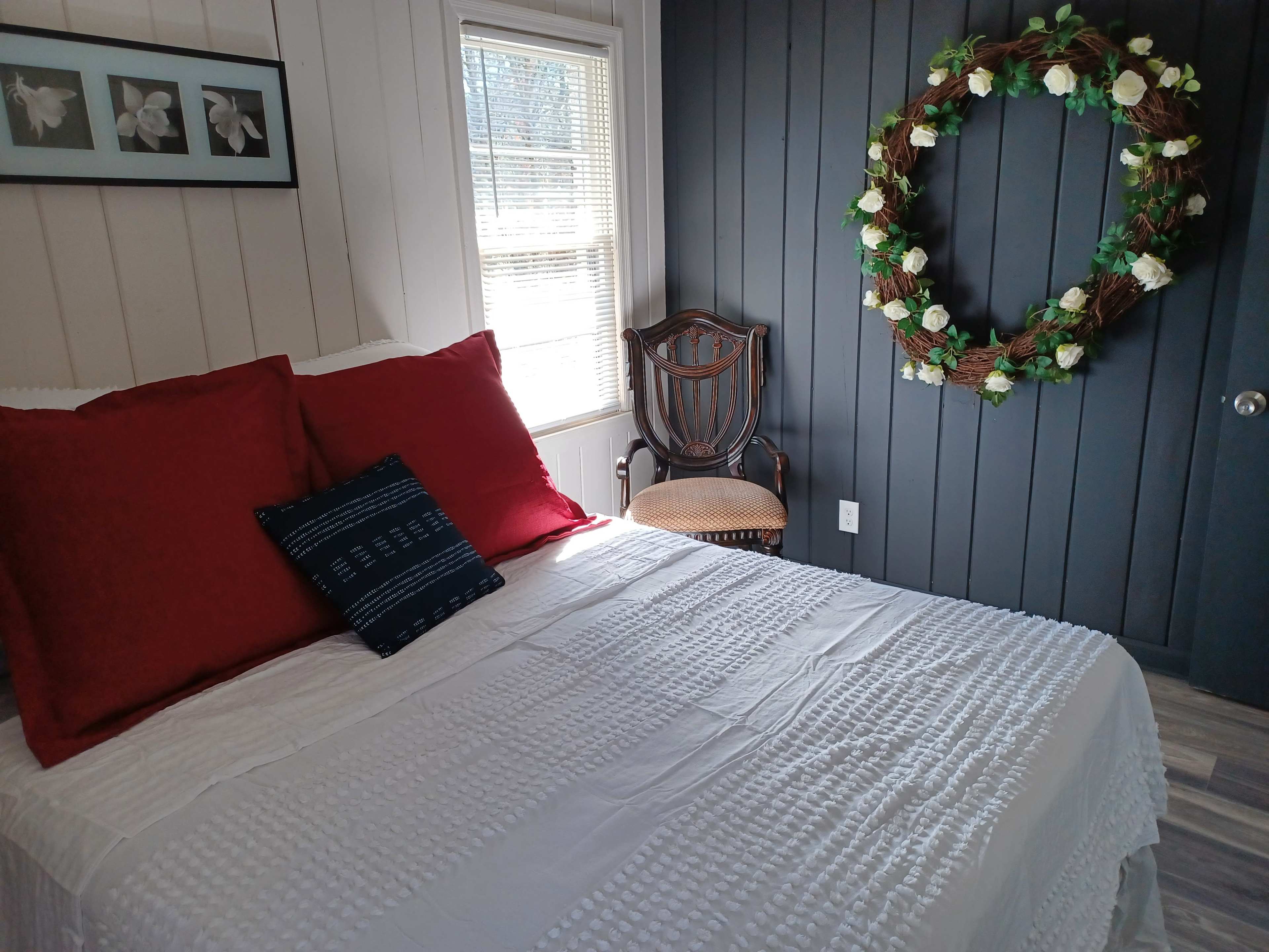 The room features a bed with a white quilt and red pillows, alongside a decorative wreath on a dark wall and a chair in the corner.