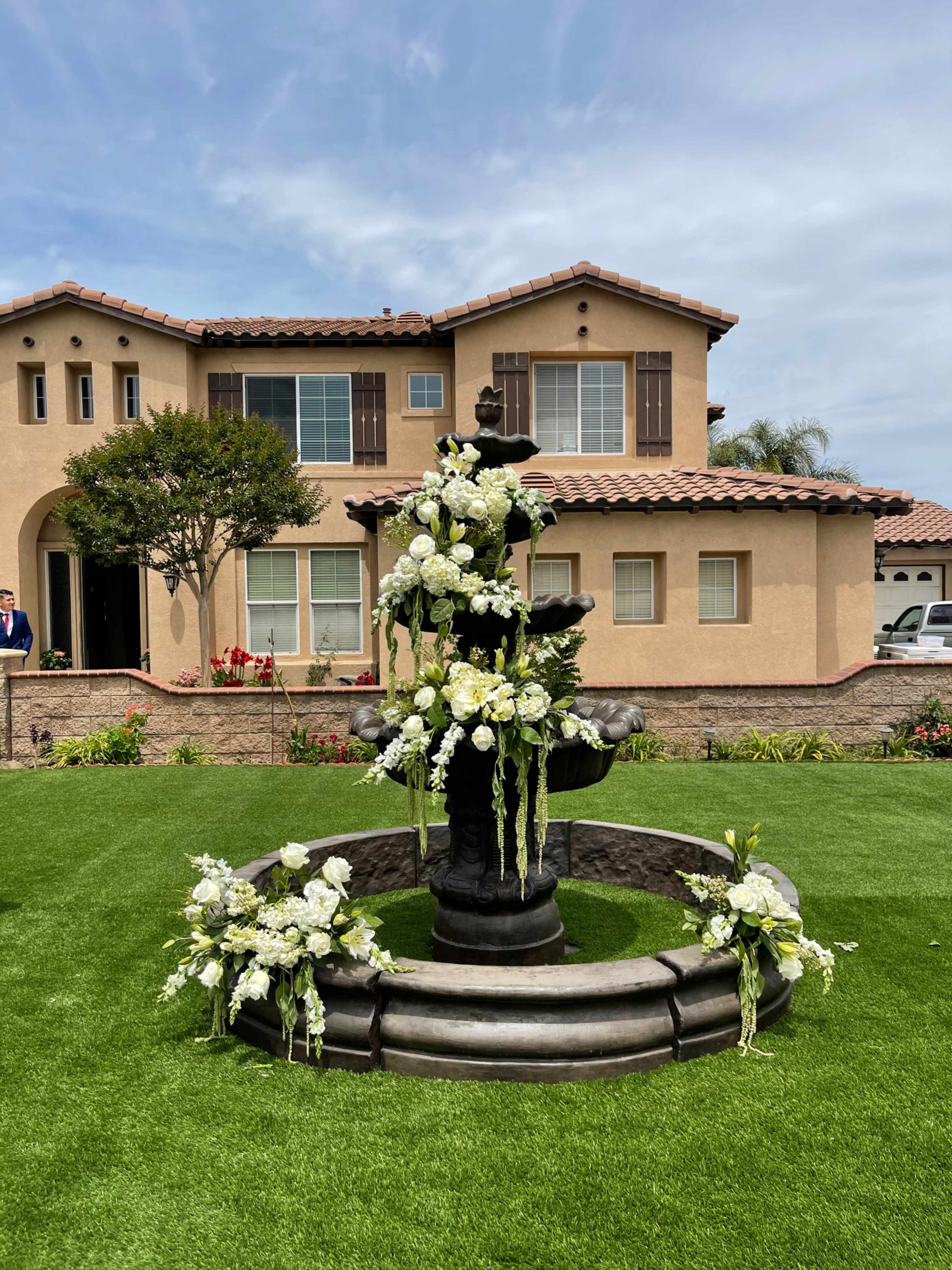 A decorative fountain adorned with white flowers is situated in the front yard of a two-story house.