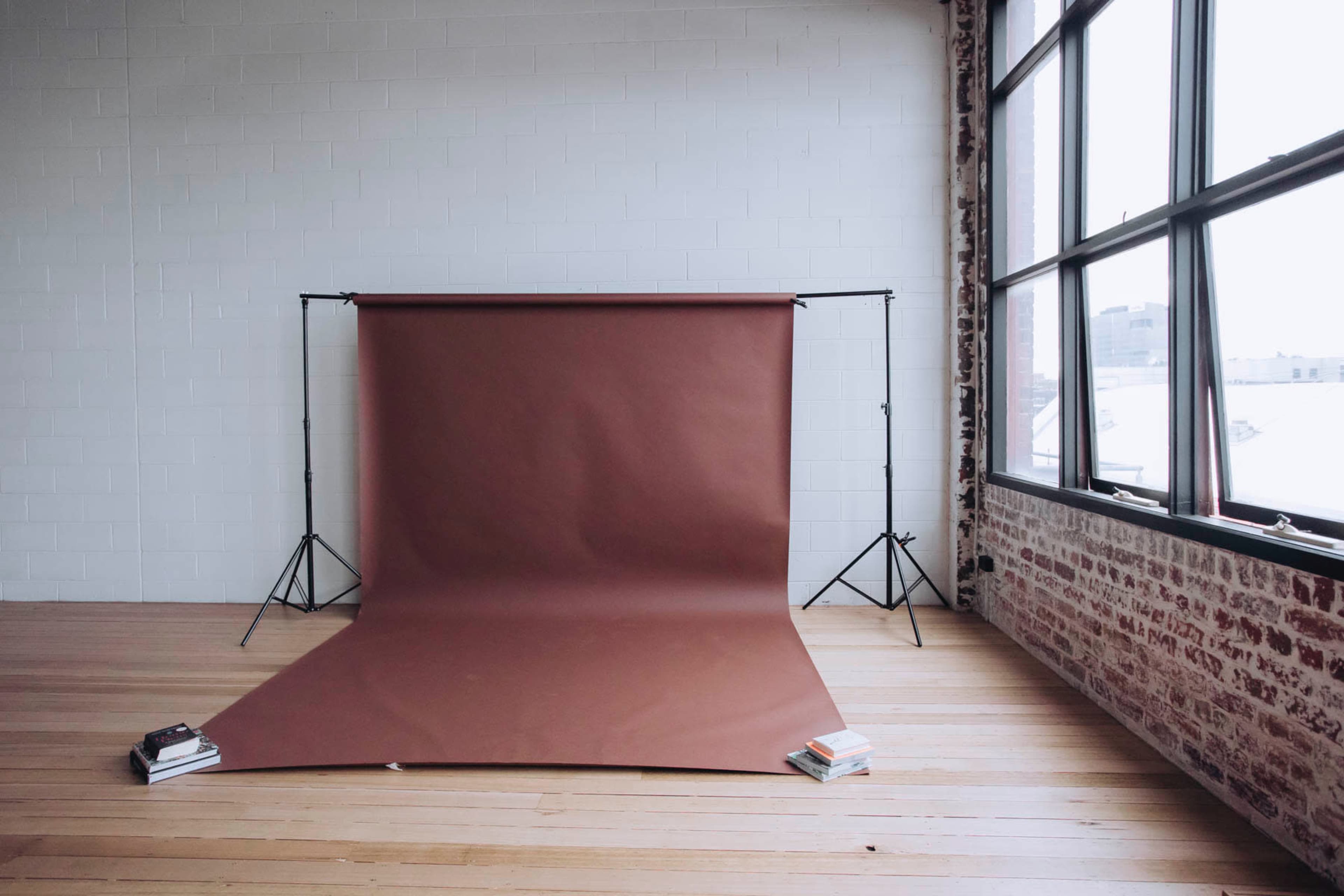 A photography backdrop stands on a frame in a spacious room with large windows, and two stacks of books are placed on the floor at the base.