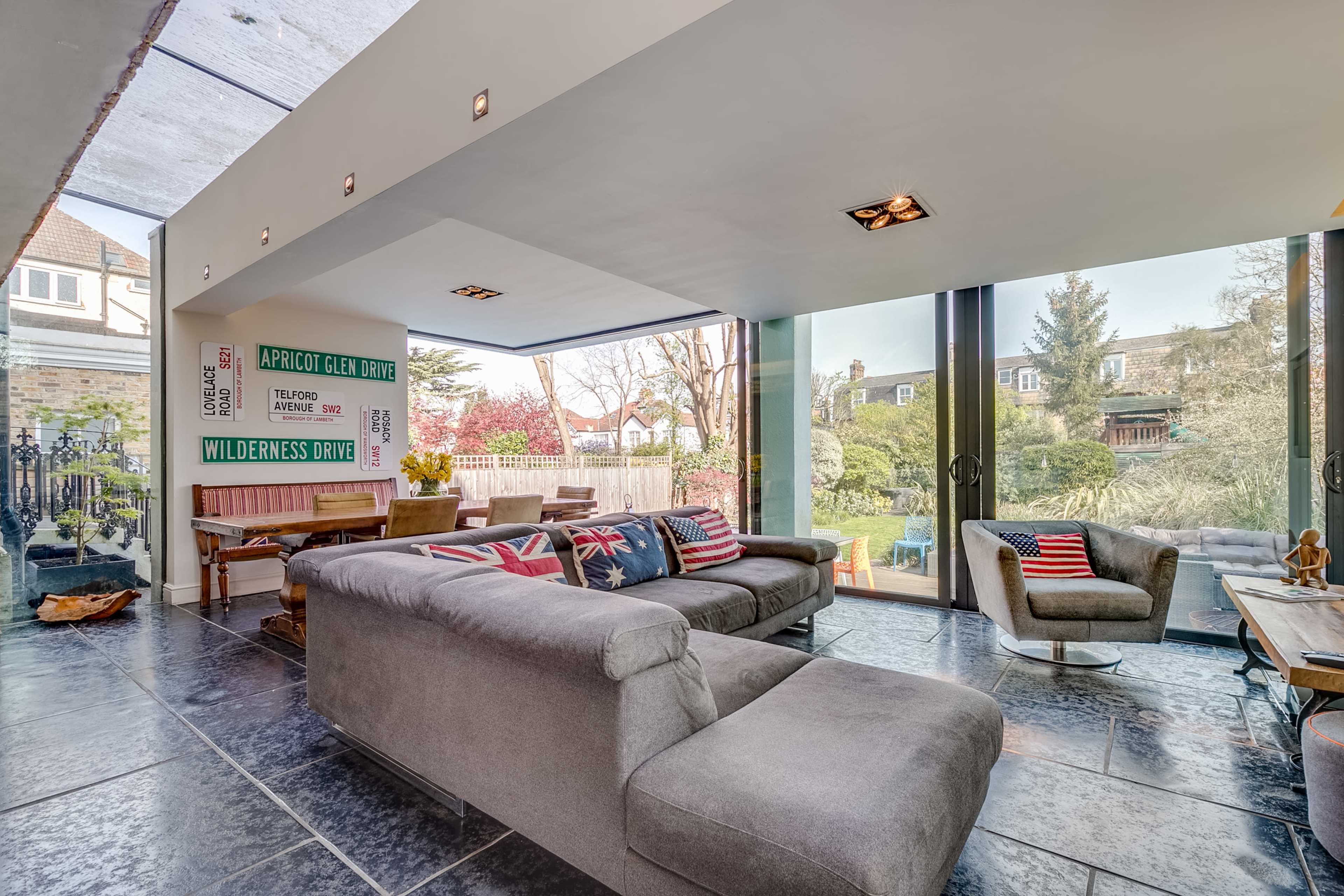 A modern living room with large glass windows overlooking a garden, featuring gray sofas and a wooden dining table.
