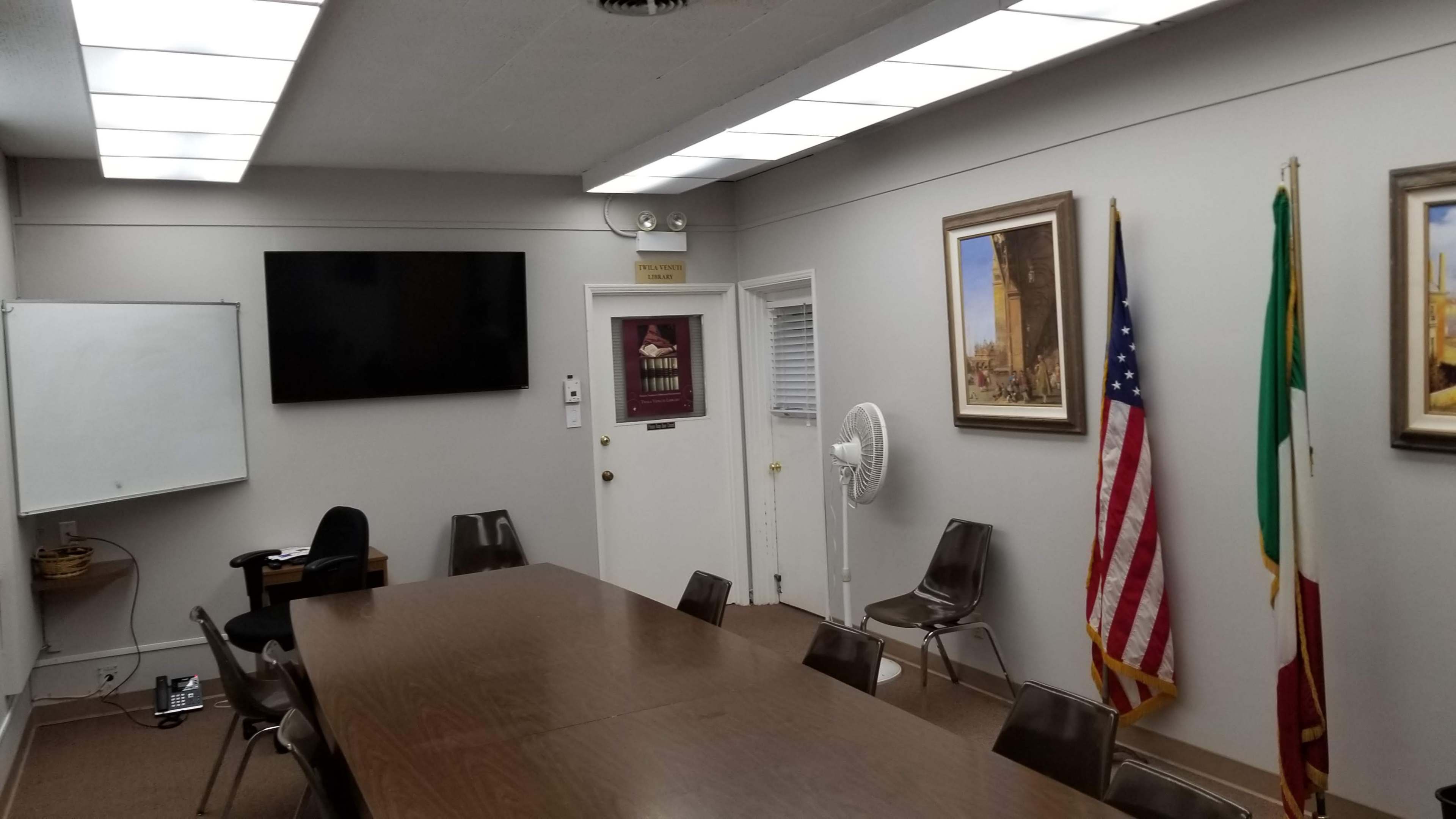 A conference room features a long wooden table surrounded by chairs, a large wall-mounted screen, and framed pictures, with flags displayed in the corner.