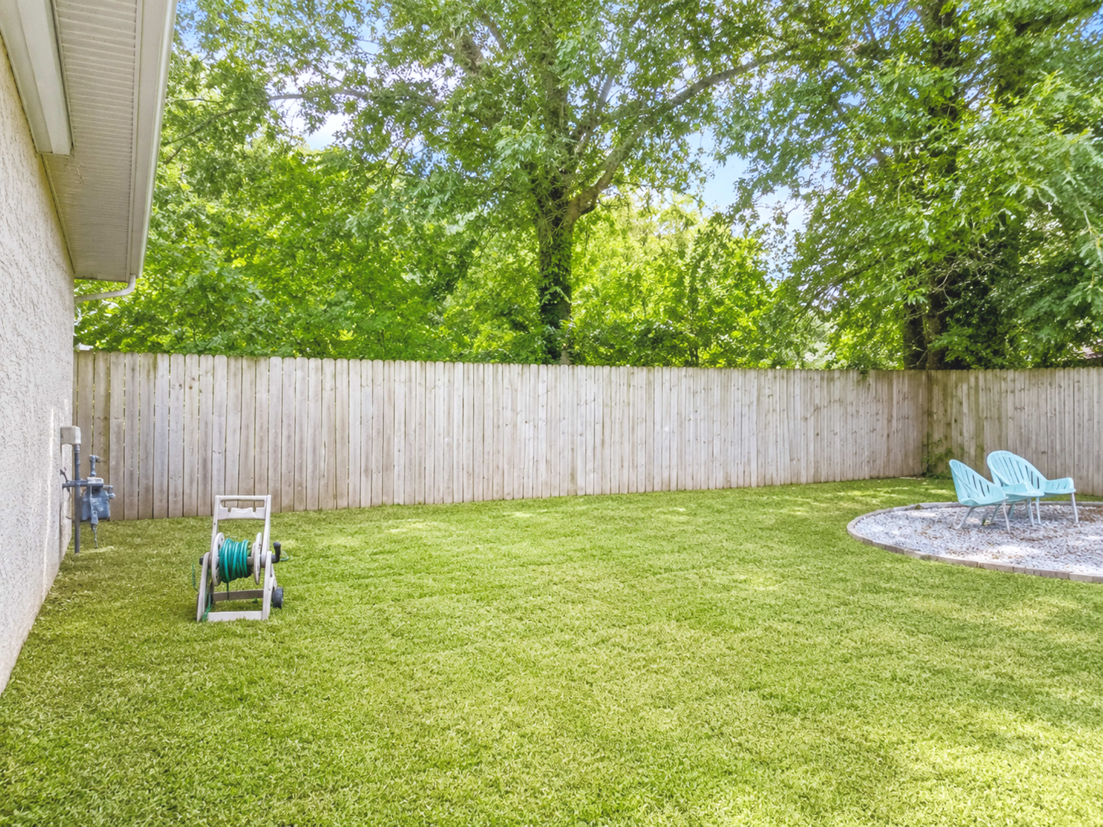 A neatly maintained backyard features a grassy area, a wooden fence, a hose reel, and light blue patio chairs.