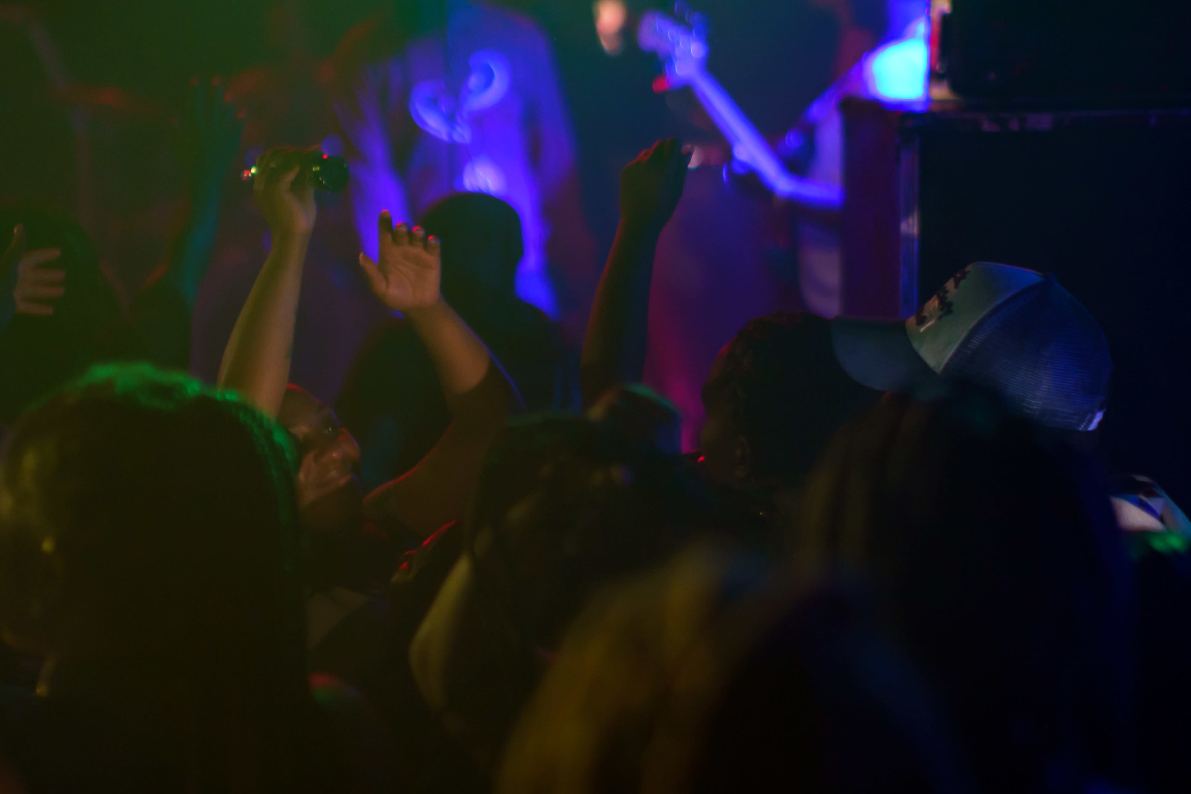 A crowded music venue is filled with fans raising their arms in excitement under colorful stage lights.