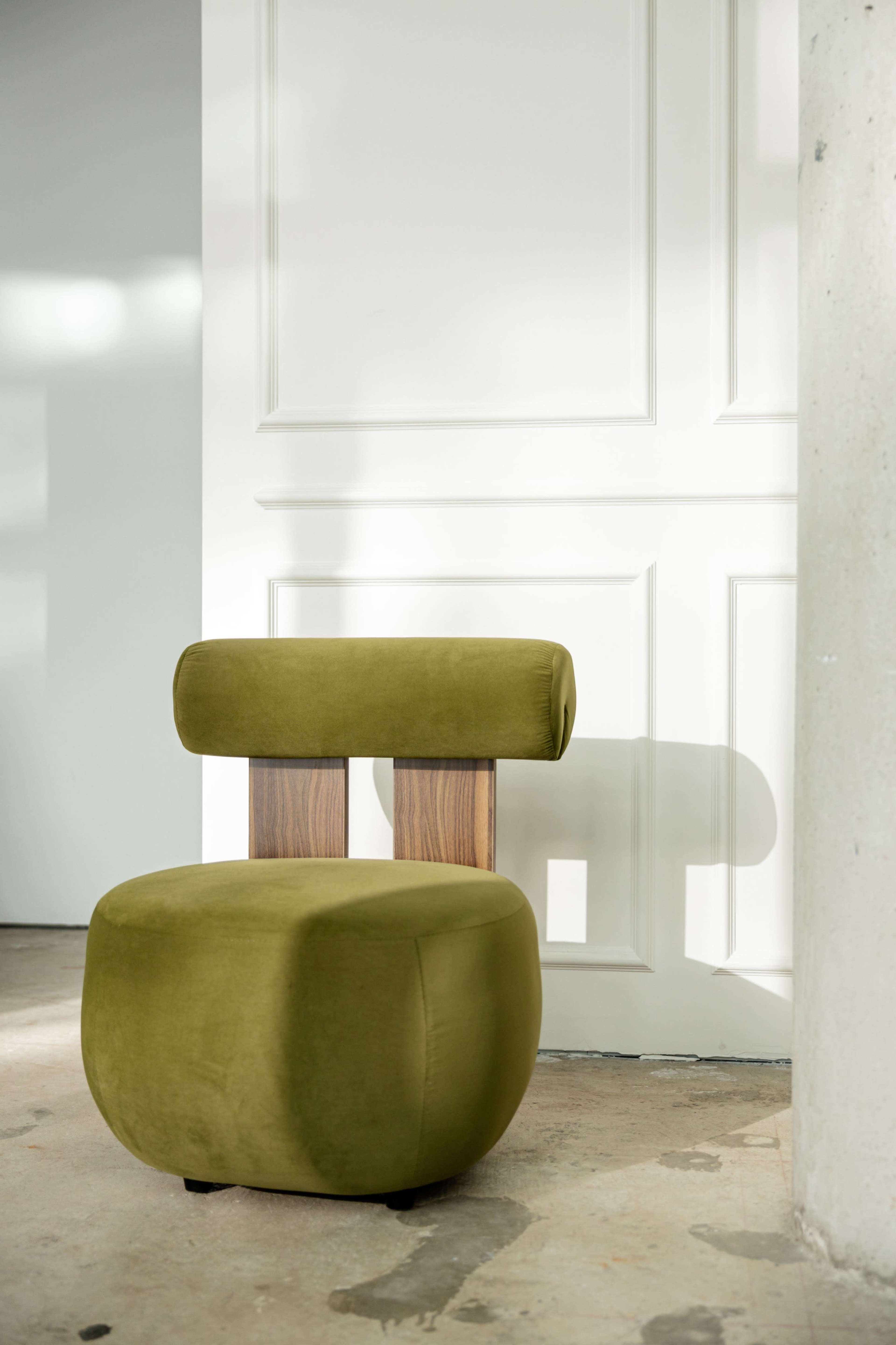 A green upholstered chair with a round seat and a backrest sits next to a white wall with paneling.