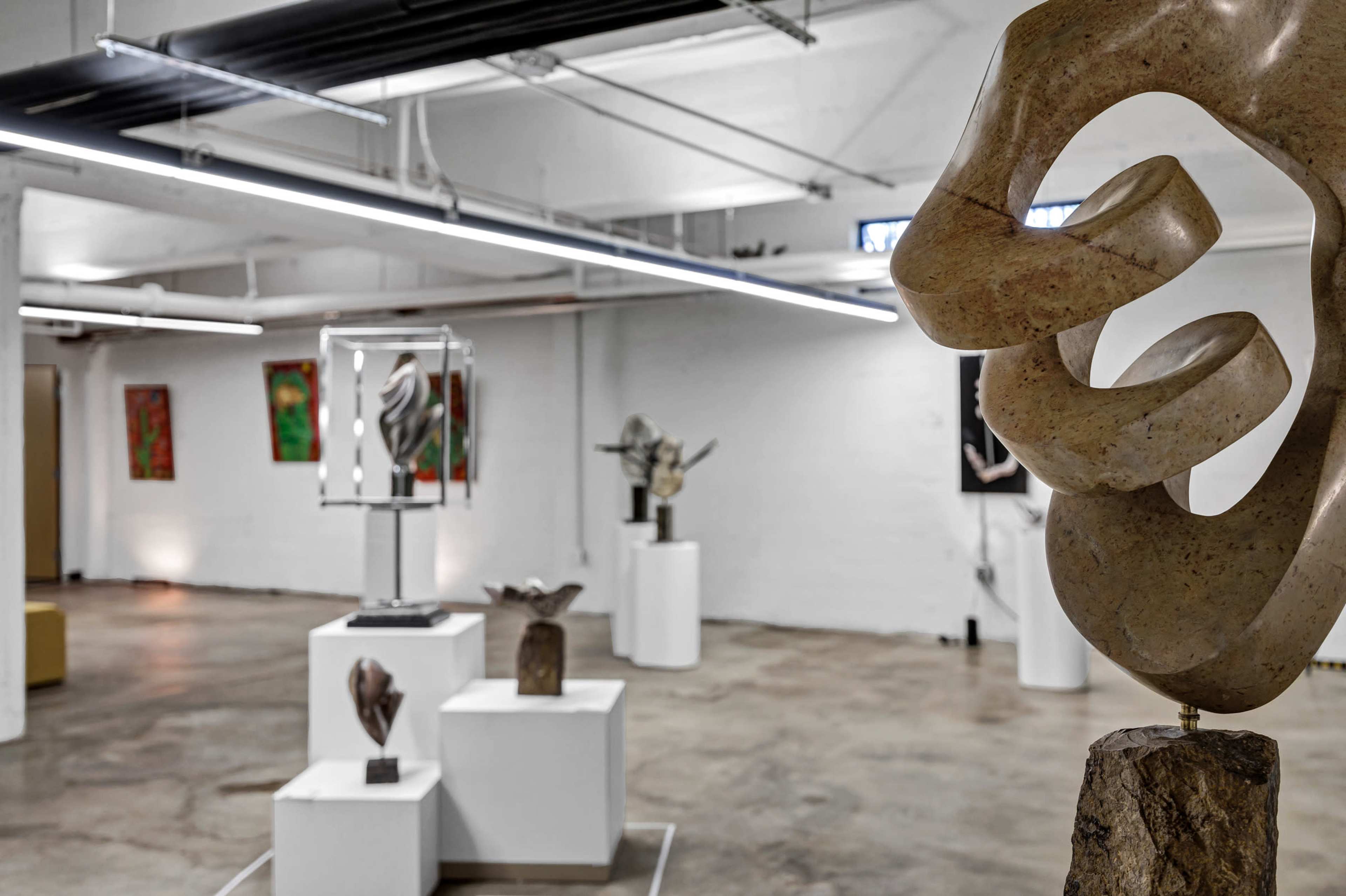 The image shows an art gallery featuring various sculptures displayed on pedestals, with colorful abstract paintings on the walls.