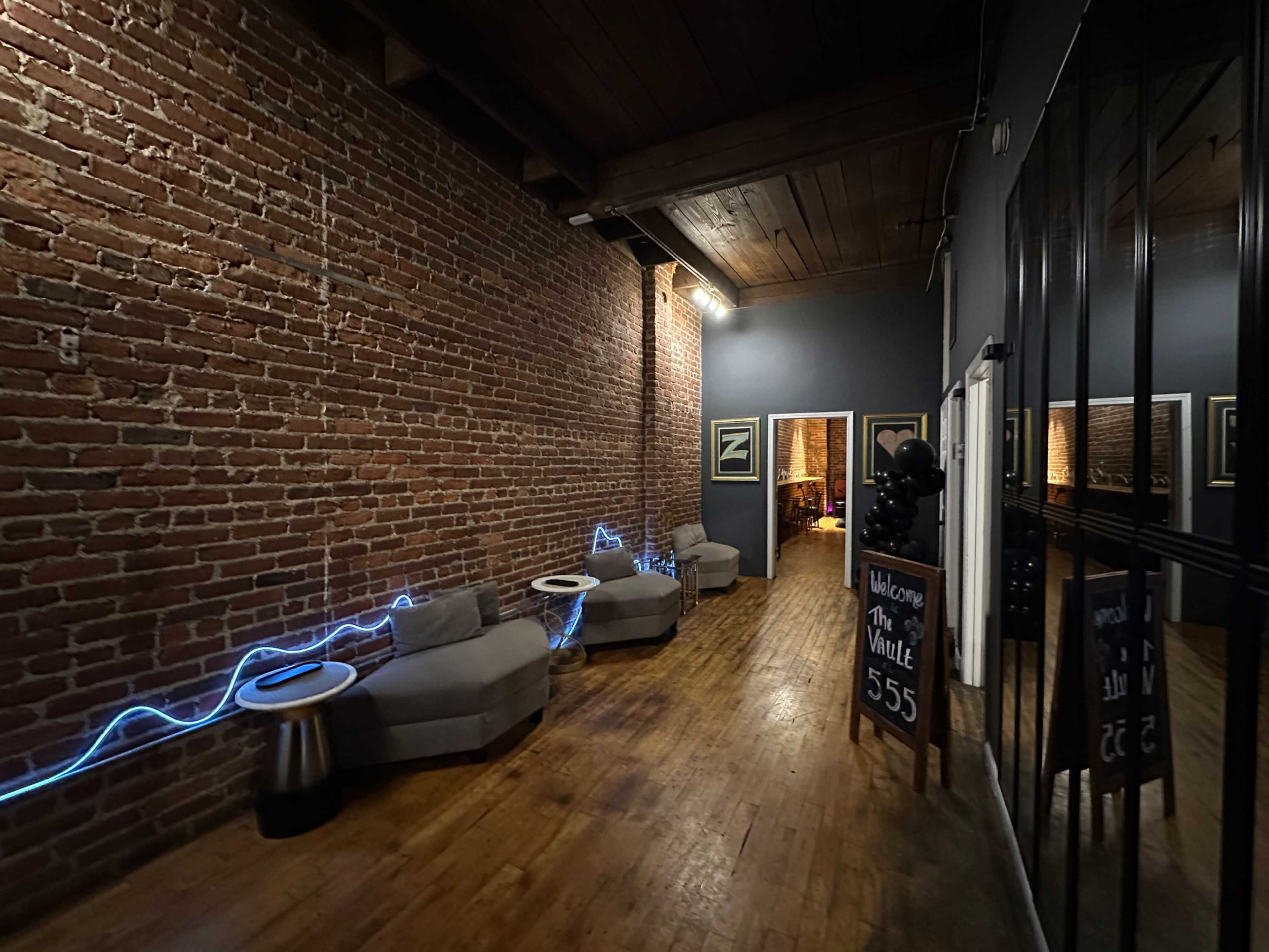 The image shows a narrow corridor with exposed brick walls, wooden flooring, and modern seating arrangements illuminated by blue neon lighting.