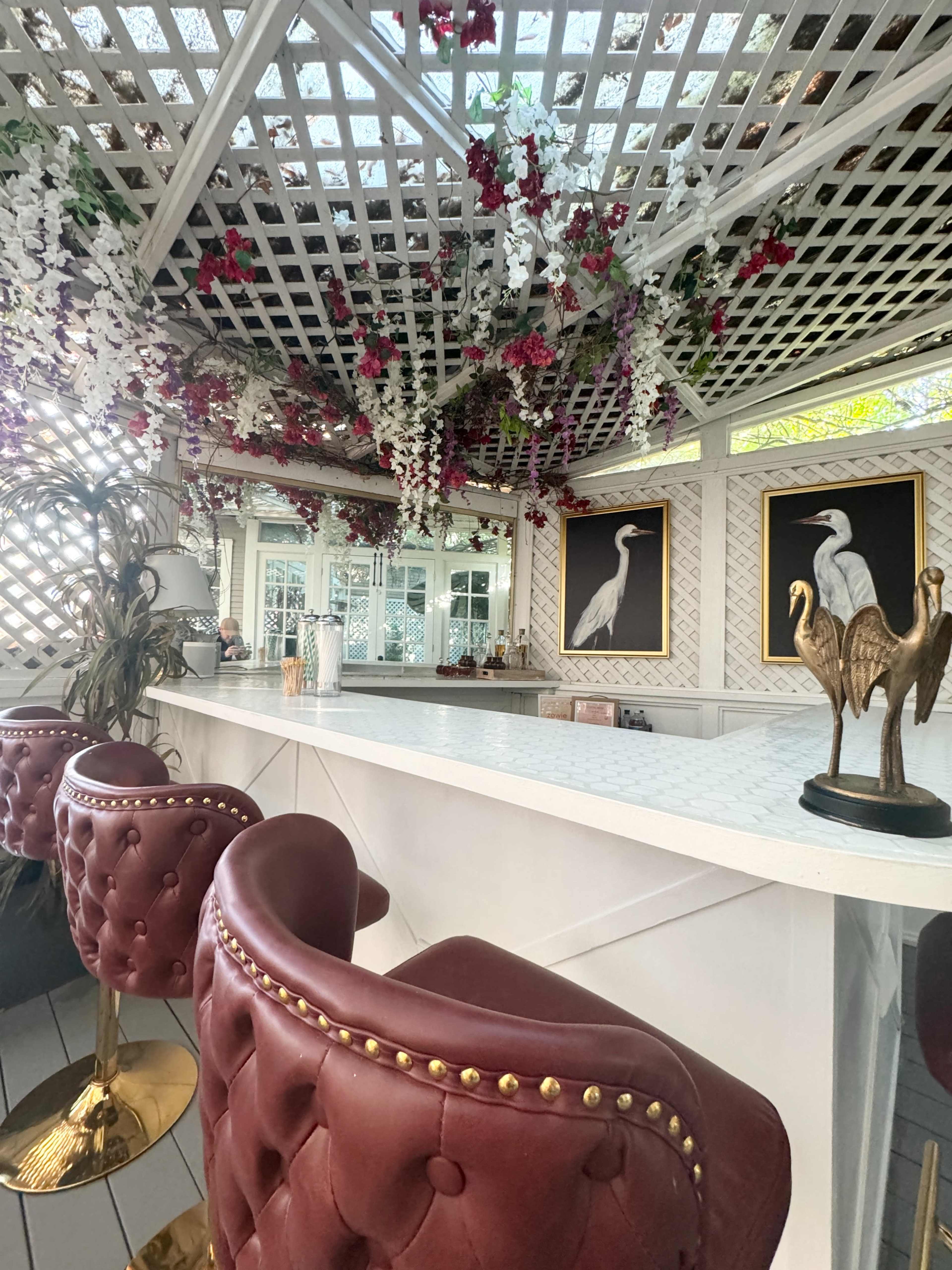 The image shows a bar area with red upholstered stools, a white countertop, and a ceiling lattice adorned with hanging flowers and artwork of birds.