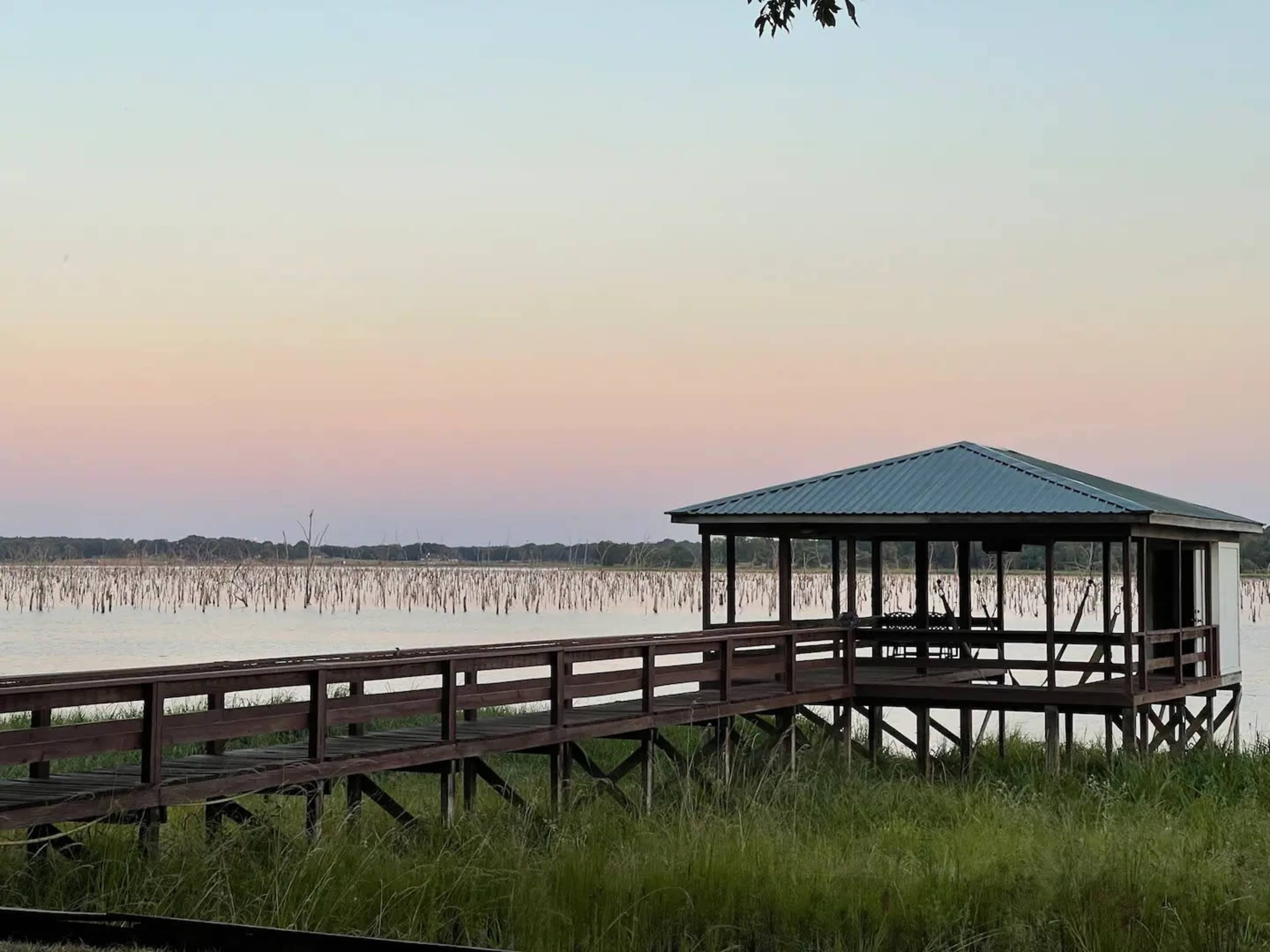 A wooden pier extends over a calm lake, with a pavilion at the end and reeds visible in the water.