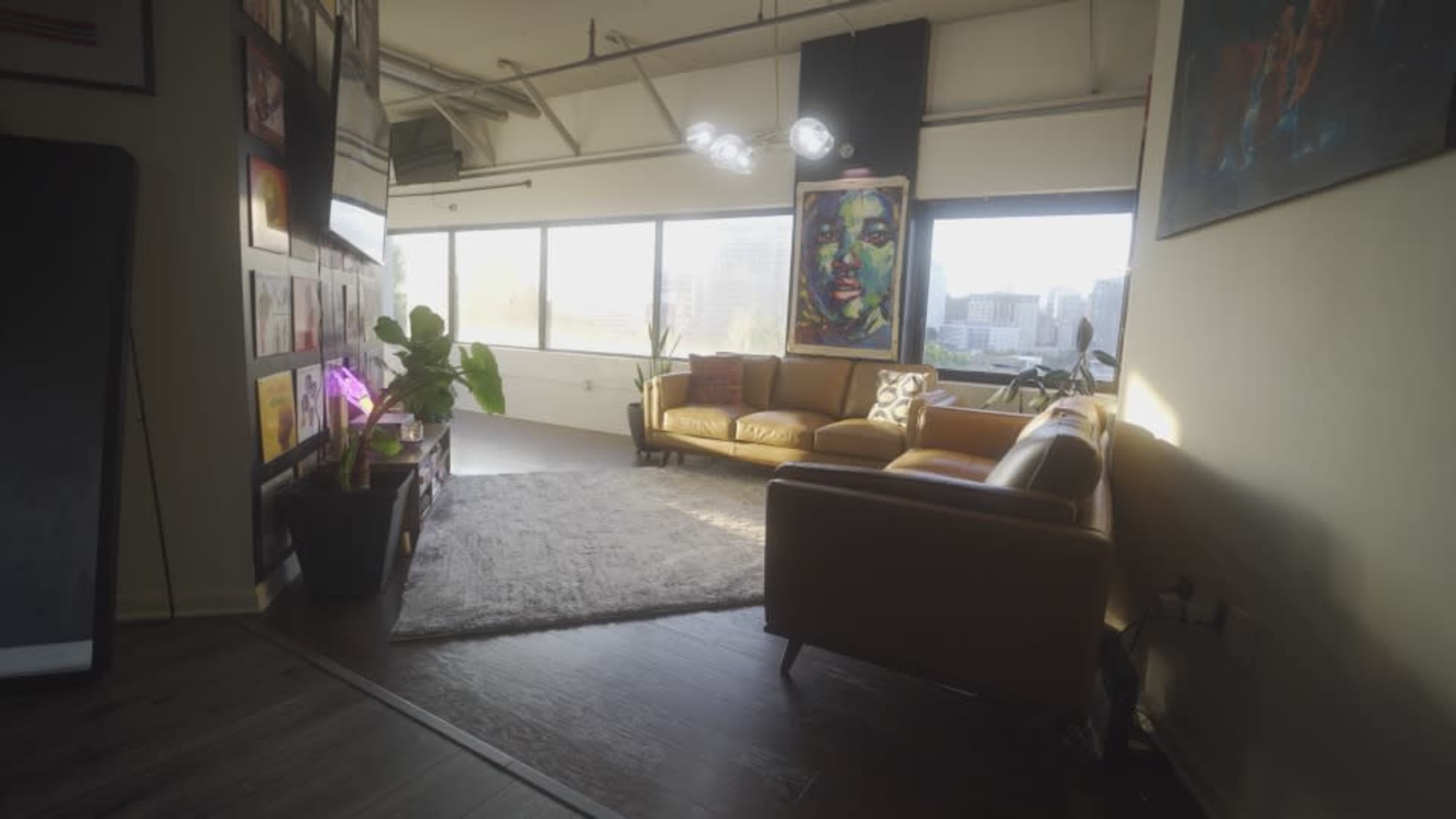 The image shows a modern, light-filled room featuring a cozy seating area with a brown leather sofa, a large rug, and decorative artwork on the walls.