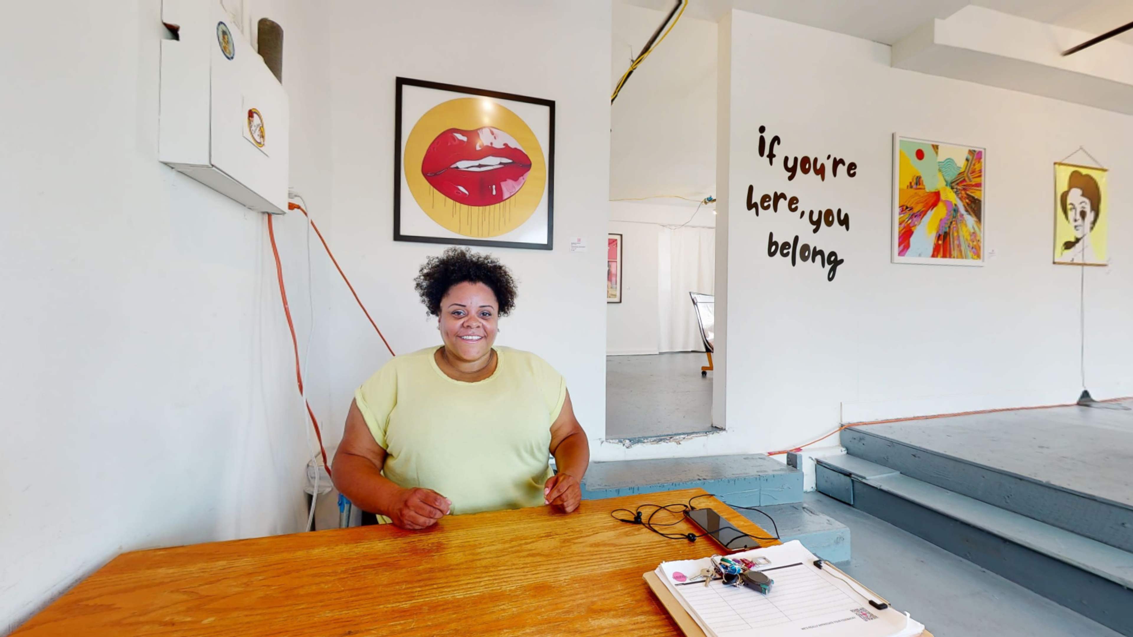 A woman sits at a wooden desk in a bright, minimalist room with colorful artwork on the walls and a motivational phrase displayed nearby.