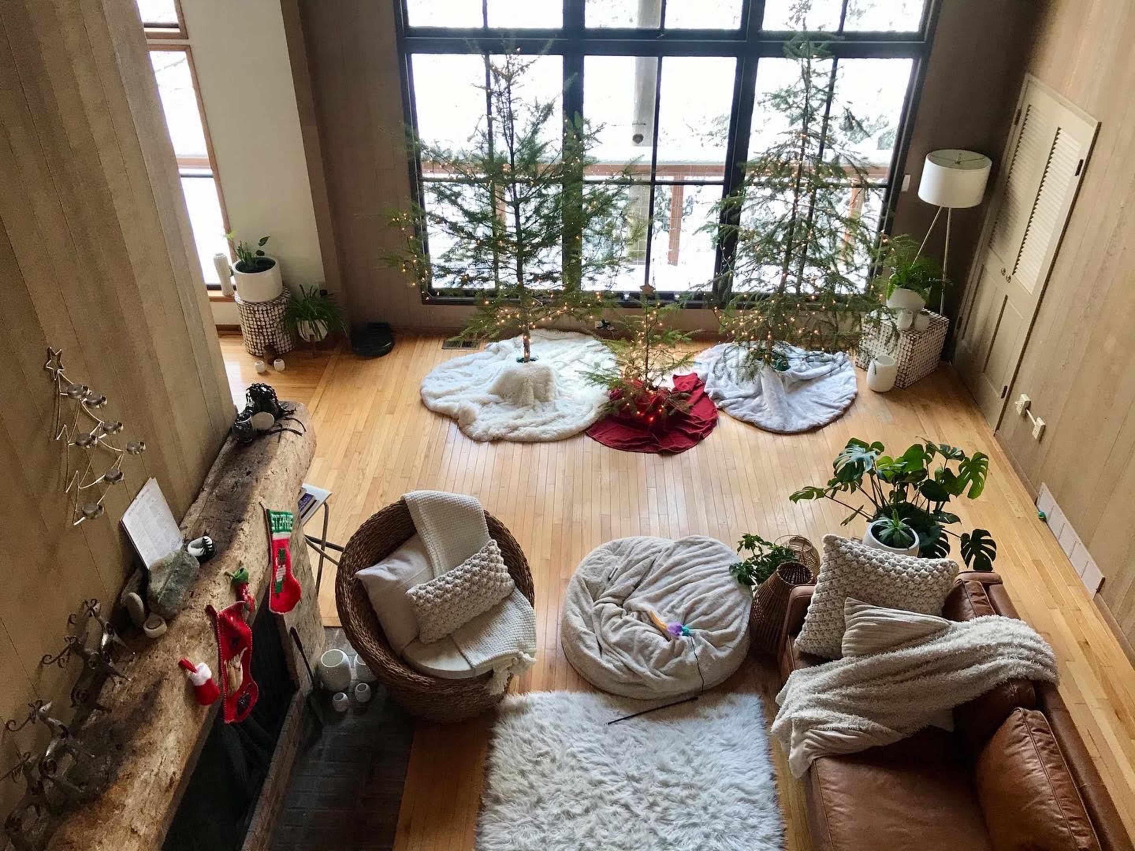 A spacious living room with two small Christmas trees on either side, a cozy couch, and various decorative elements including plants and stockings hanging by the fireplace.