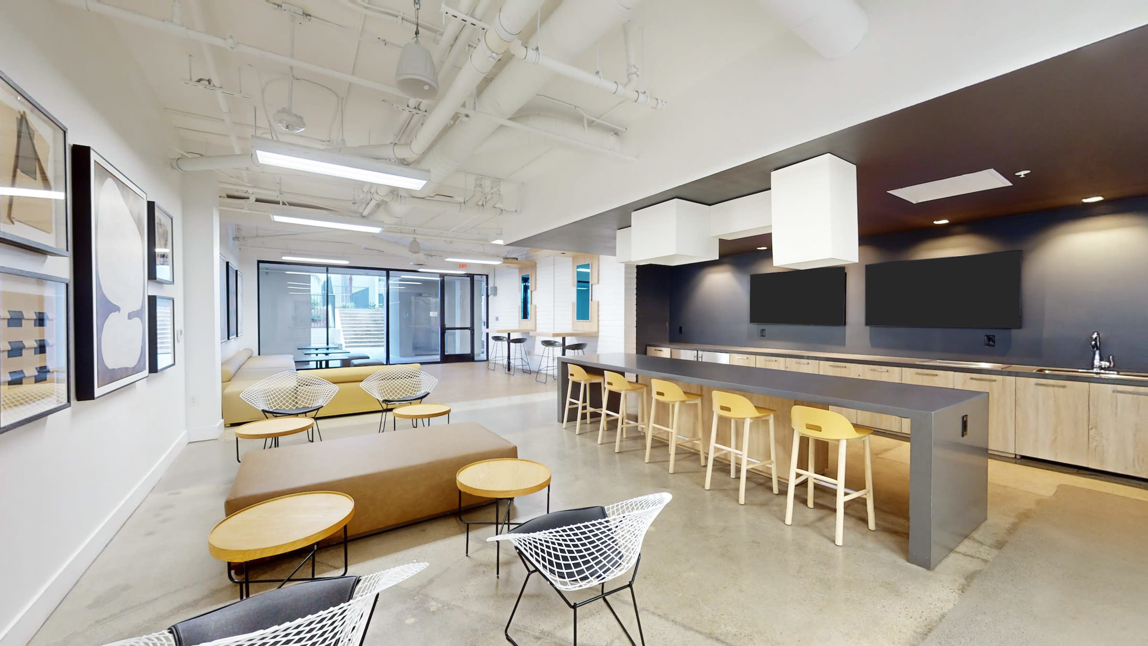 A modern office space featuring a kitchenette area with bar seating, several lounge chairs, and large windows leading to an outdoor area.
