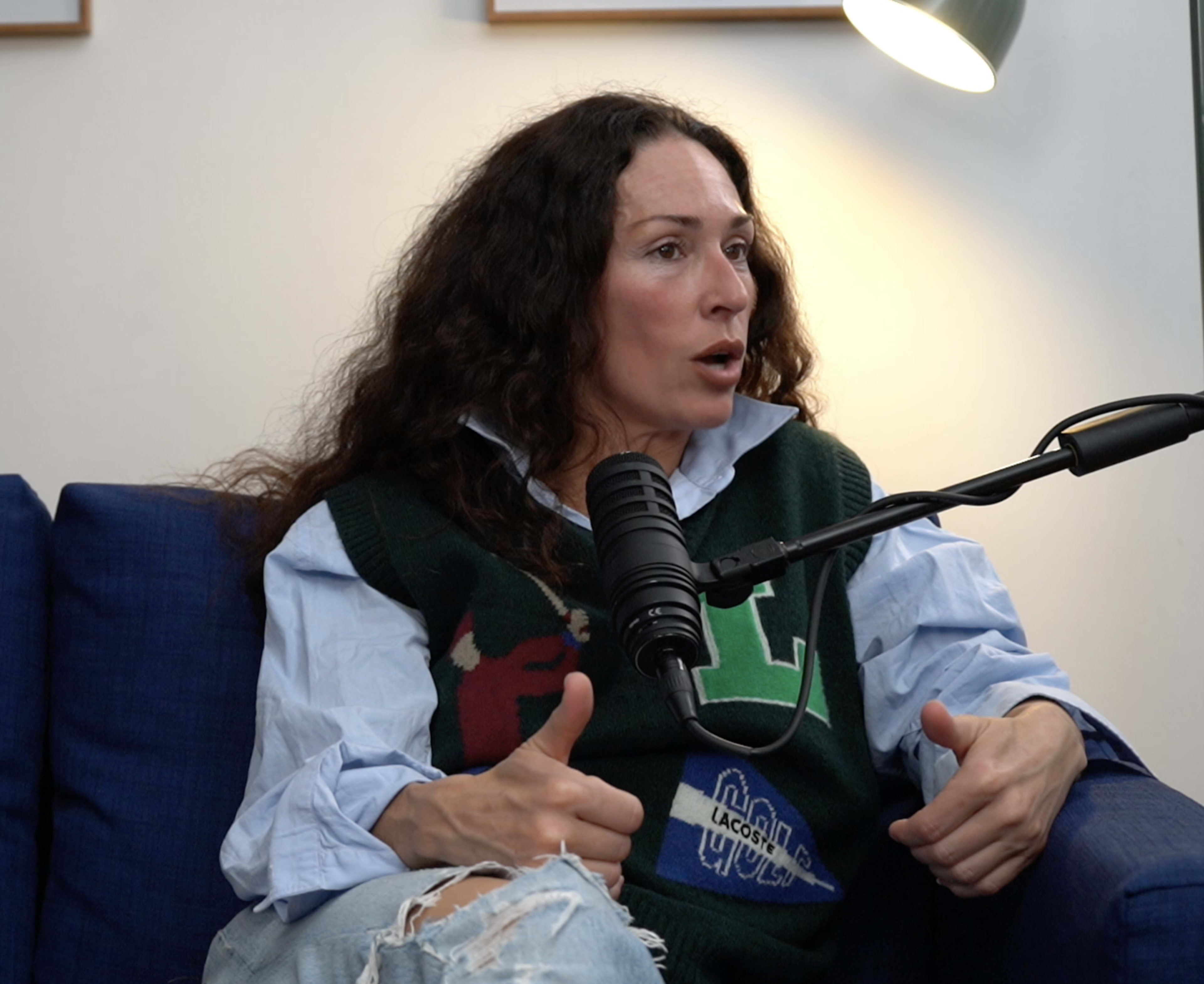 A woman with curly hair is speaking into a microphone while sitting on a blue couch, wearing a green sweater vest over a shirt with rolled-up sleeves.