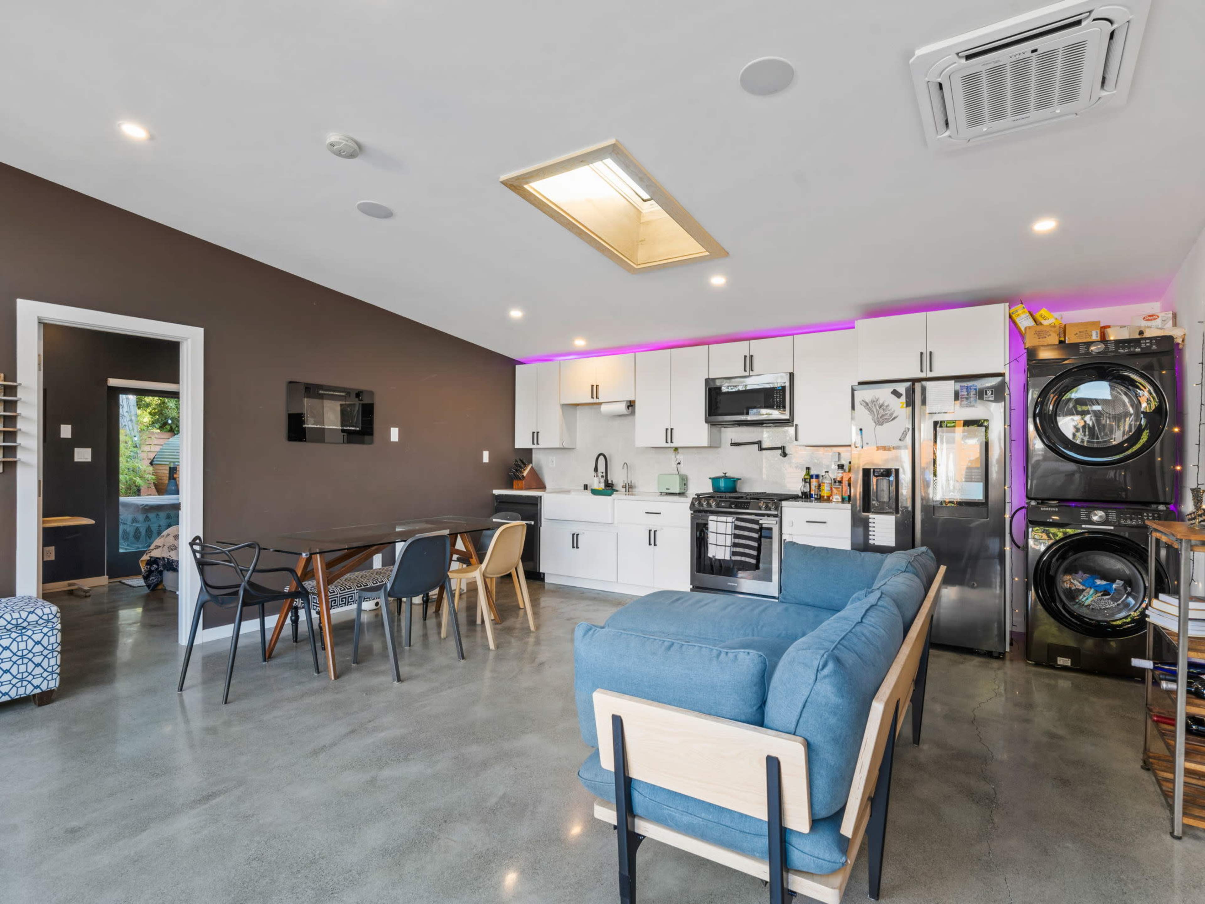 A modern kitchen and living area featuring a blue couch, a dining table, stainless steel appliances, and a stackable washer and dryer.